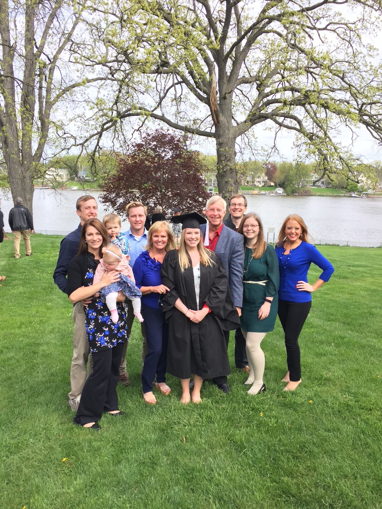 Group of adults and children posing on a grassy park by a lake; a graduate in cap and gown stands center.