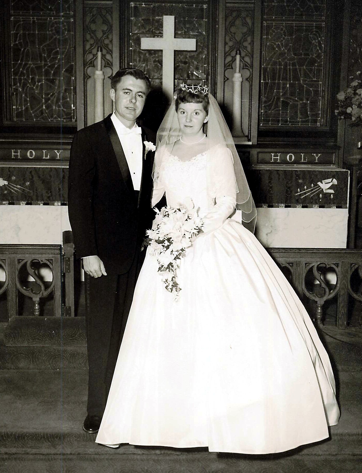 Wedding couple standing inside a church, bride in a full white gown with veil and tiara, holding a bouquet, beside a man in a black tuxedo; large cross and stained glass behind them.