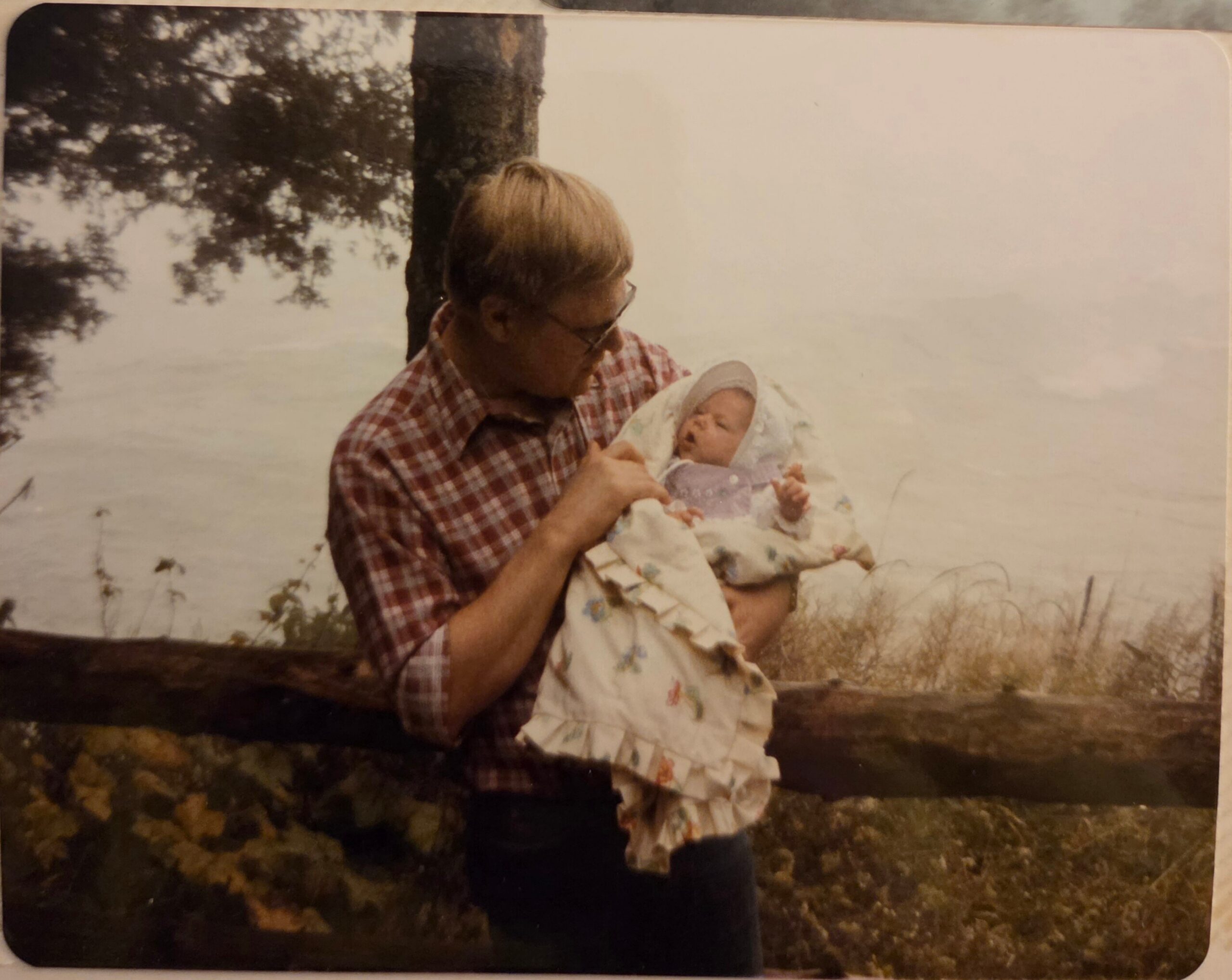 Man in a red plaid shirt holding a wrapped newborn outdoors by a tree, with a lake in the background.