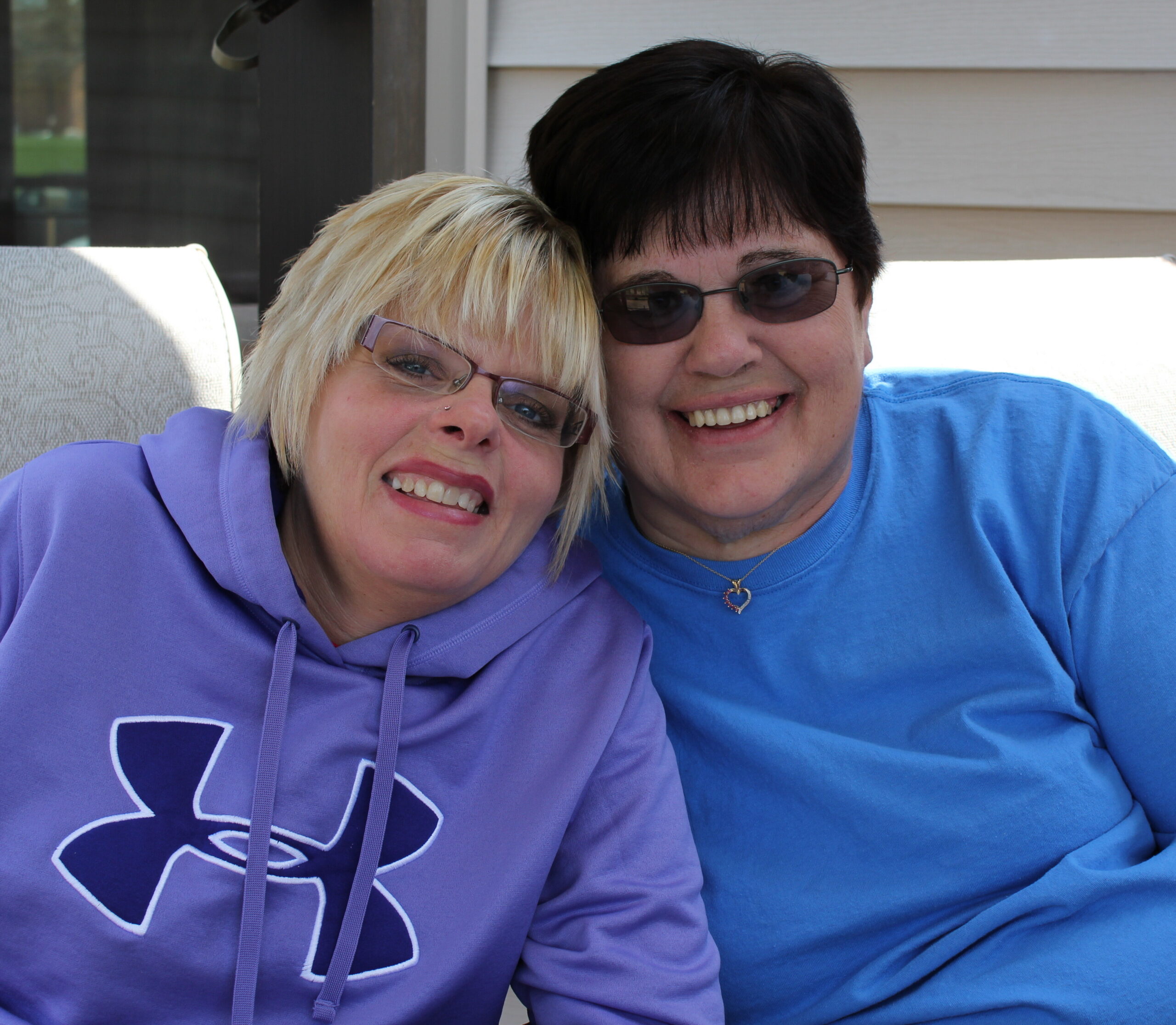 Two women lean their heads together and smile at the camera, wearing a purple hoodie and a blue shirt outdoors.