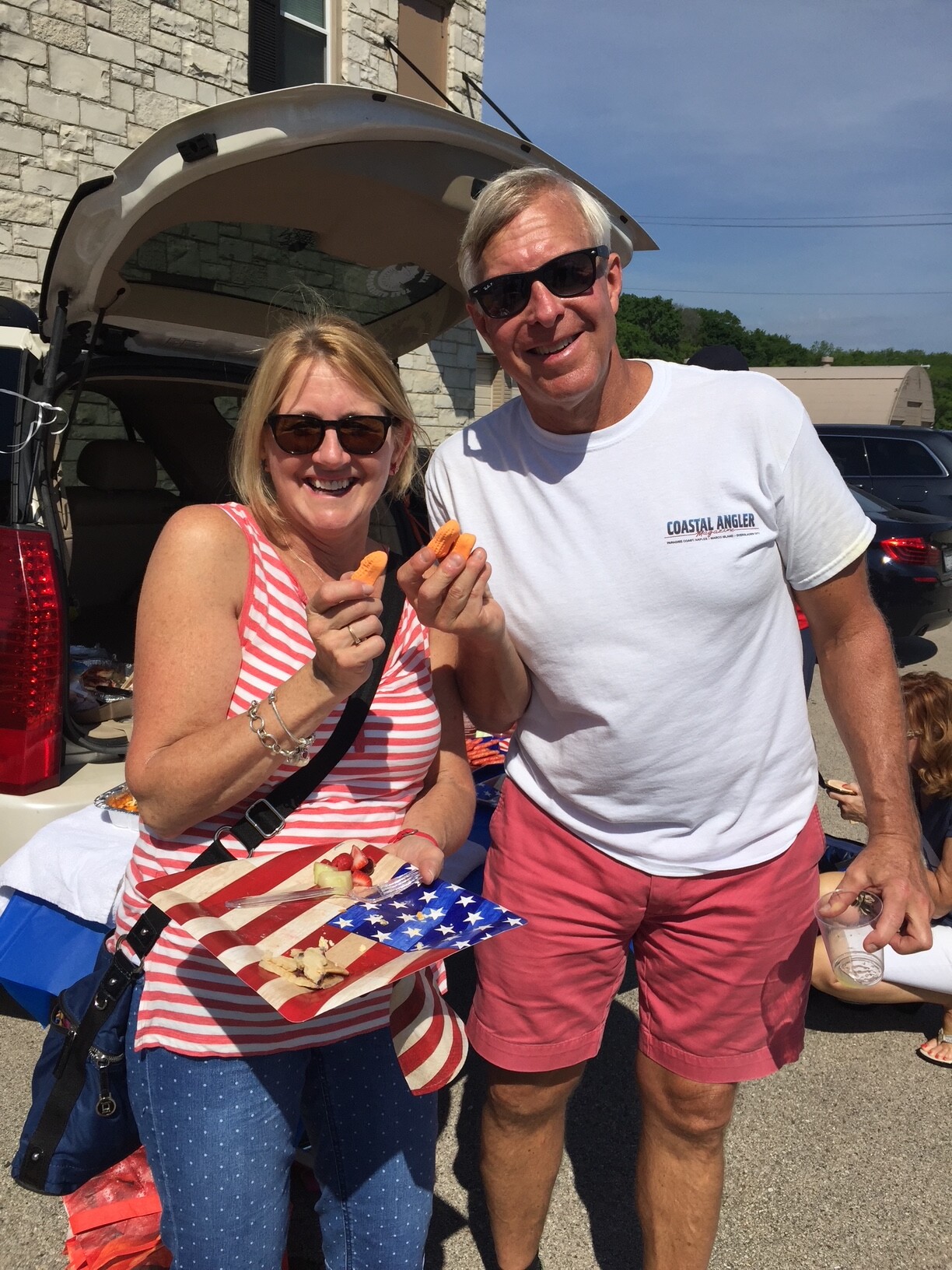 Smiling couple posing by an open car trunk, holding orange snacks on a plate with an American flag design, enjoying a sunny outdoor gathering.
