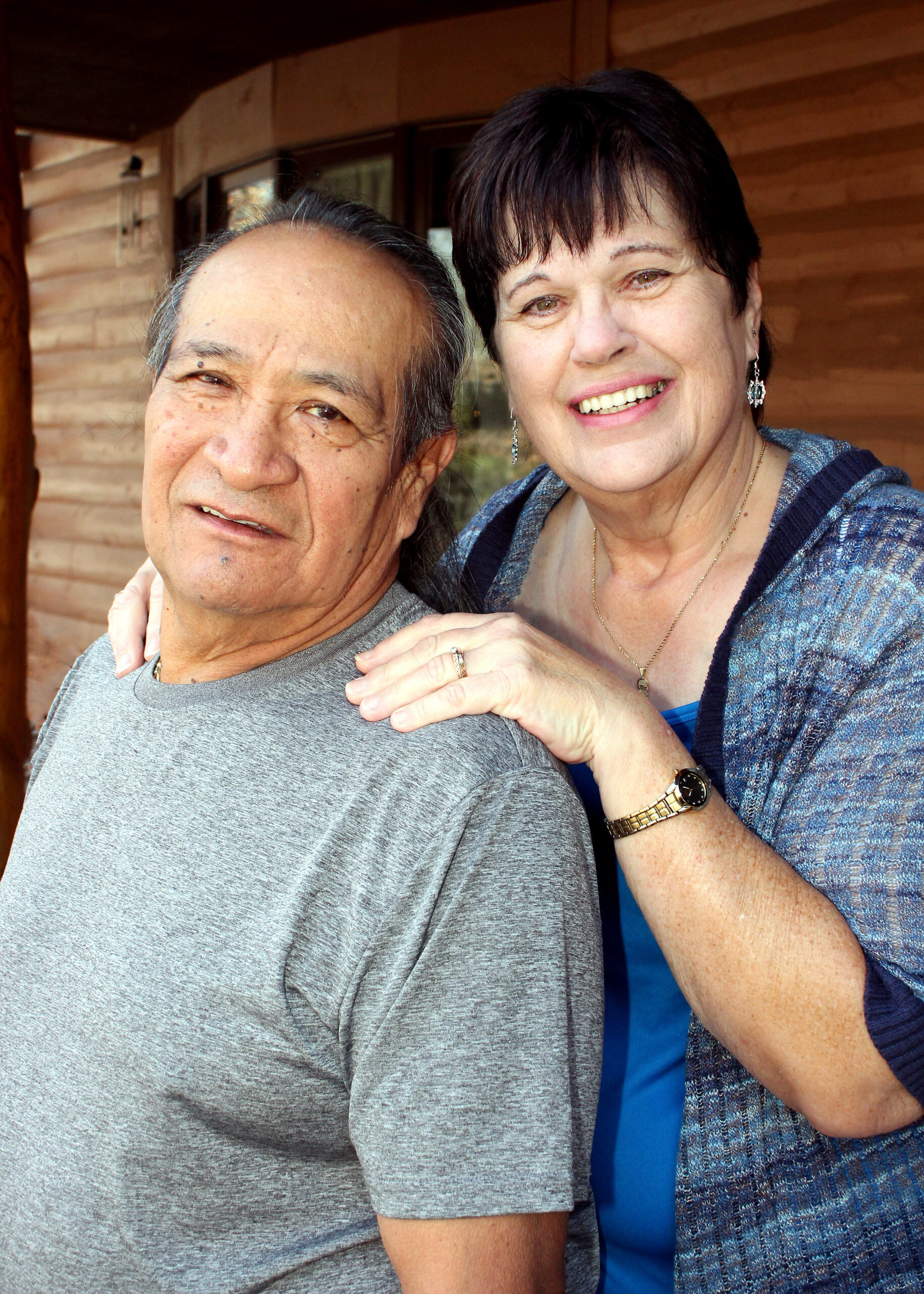 Smiling older couple posing together on a wooden porch; the woman hugs the man from behind.