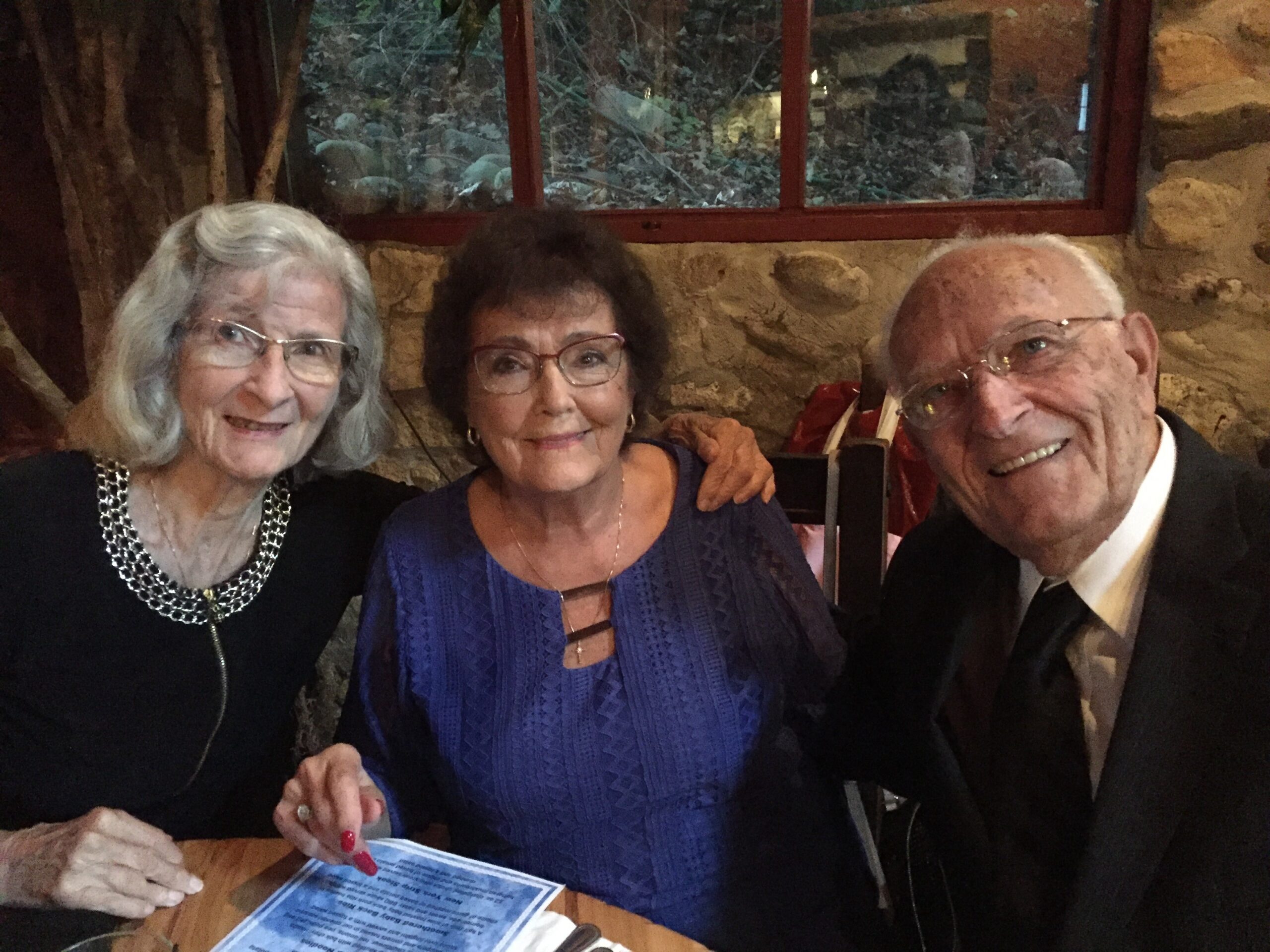 Three seniors sit together at a restaurant table, smiling at the camera with a menu in view in the foreground.