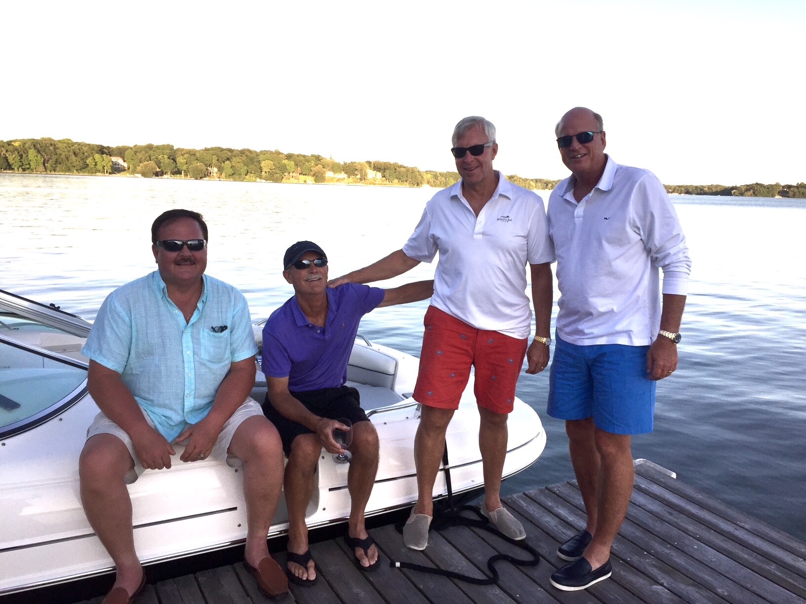 Four men in sunglasses pose on a docked white boat by a calm lake with trees in the background, daytime.