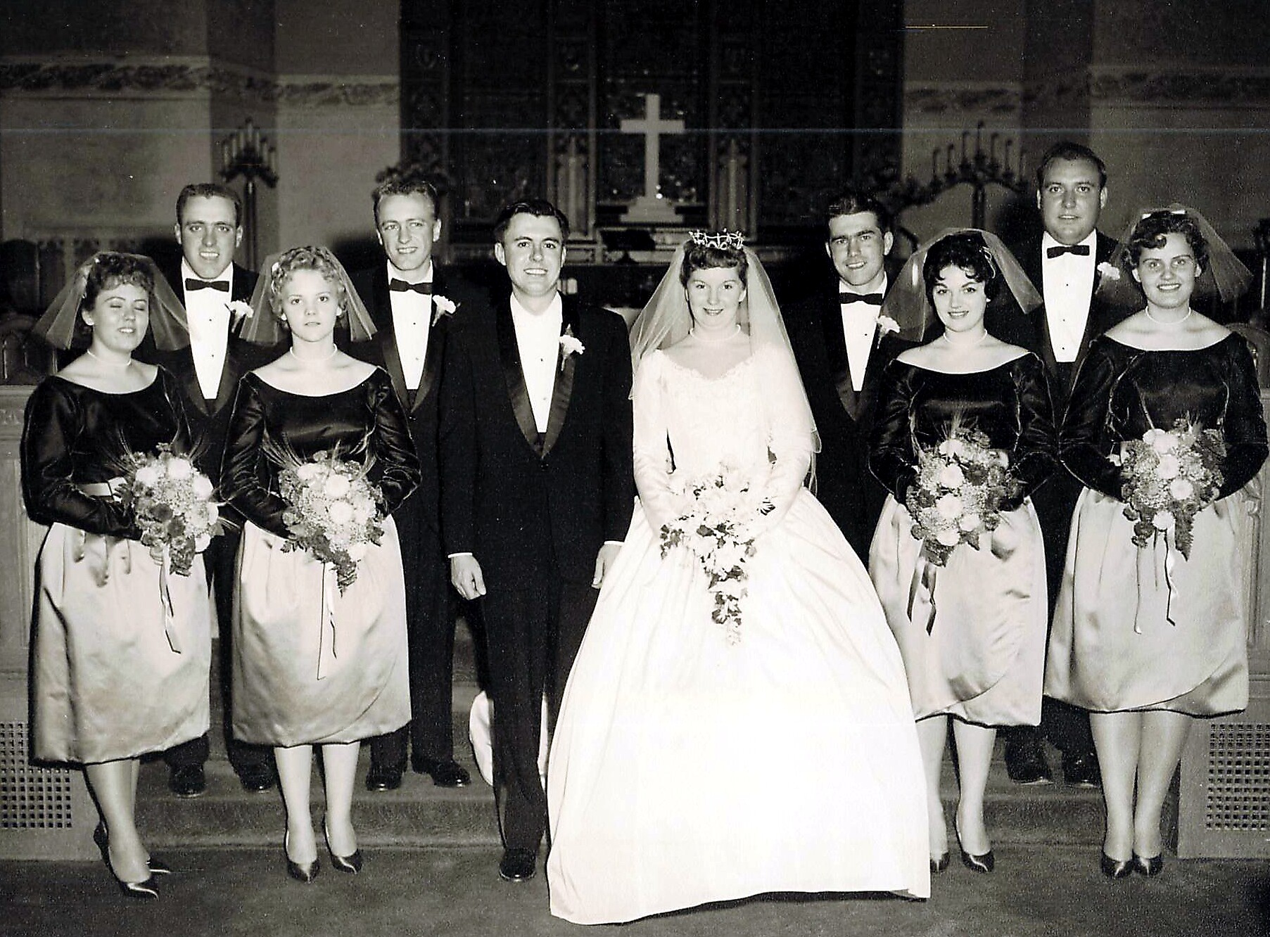 Wedding party posed in a church: bride in white gown center, groom in tuxedo, bridesmaids in black dresses, groomsmen in tuxedos, altar with cross behind.