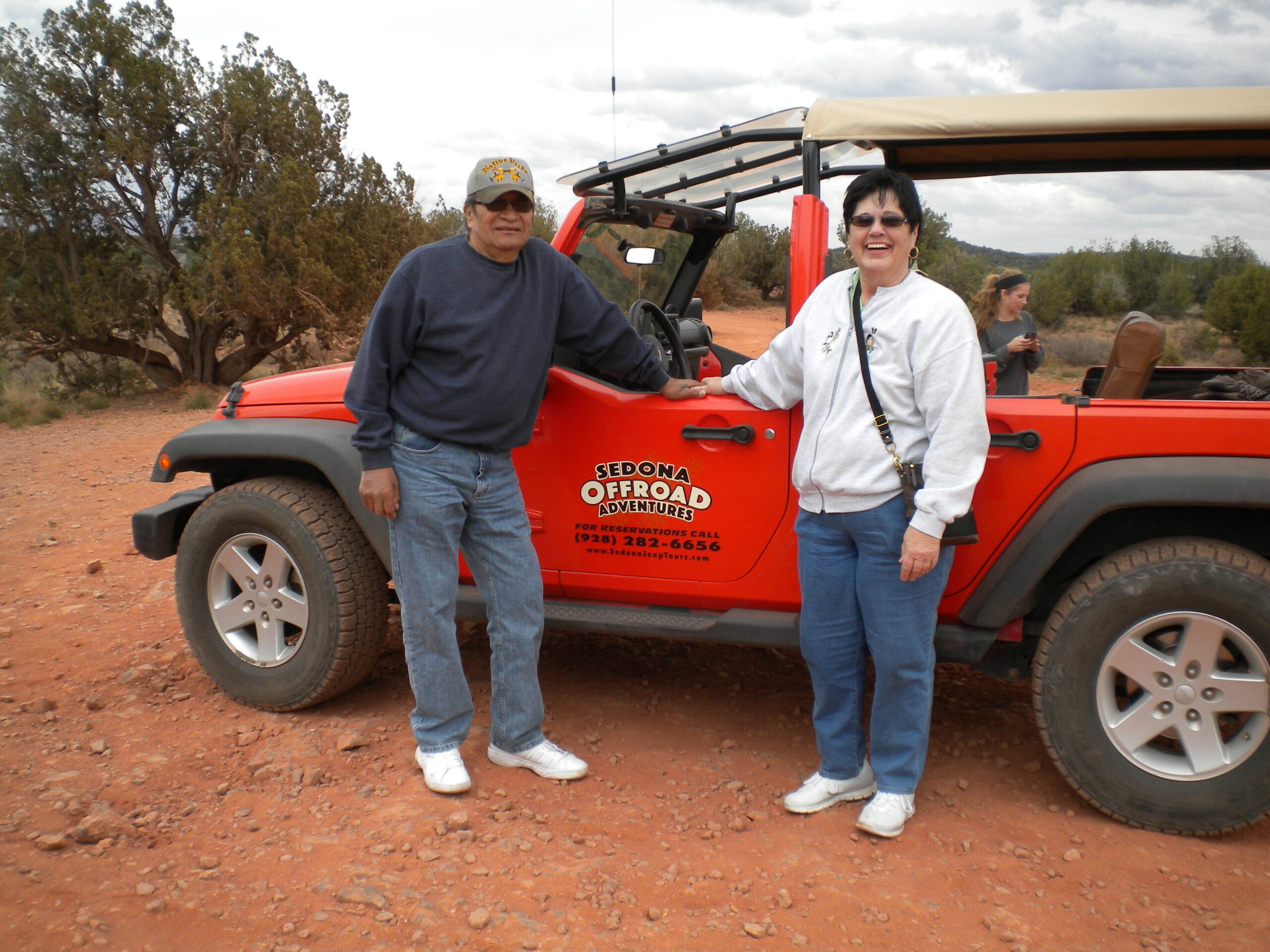 Two adults pose beside a red open-top off-road vehicle in a desert landscape, smiling at the camera (Sedona Offroad Adventures).