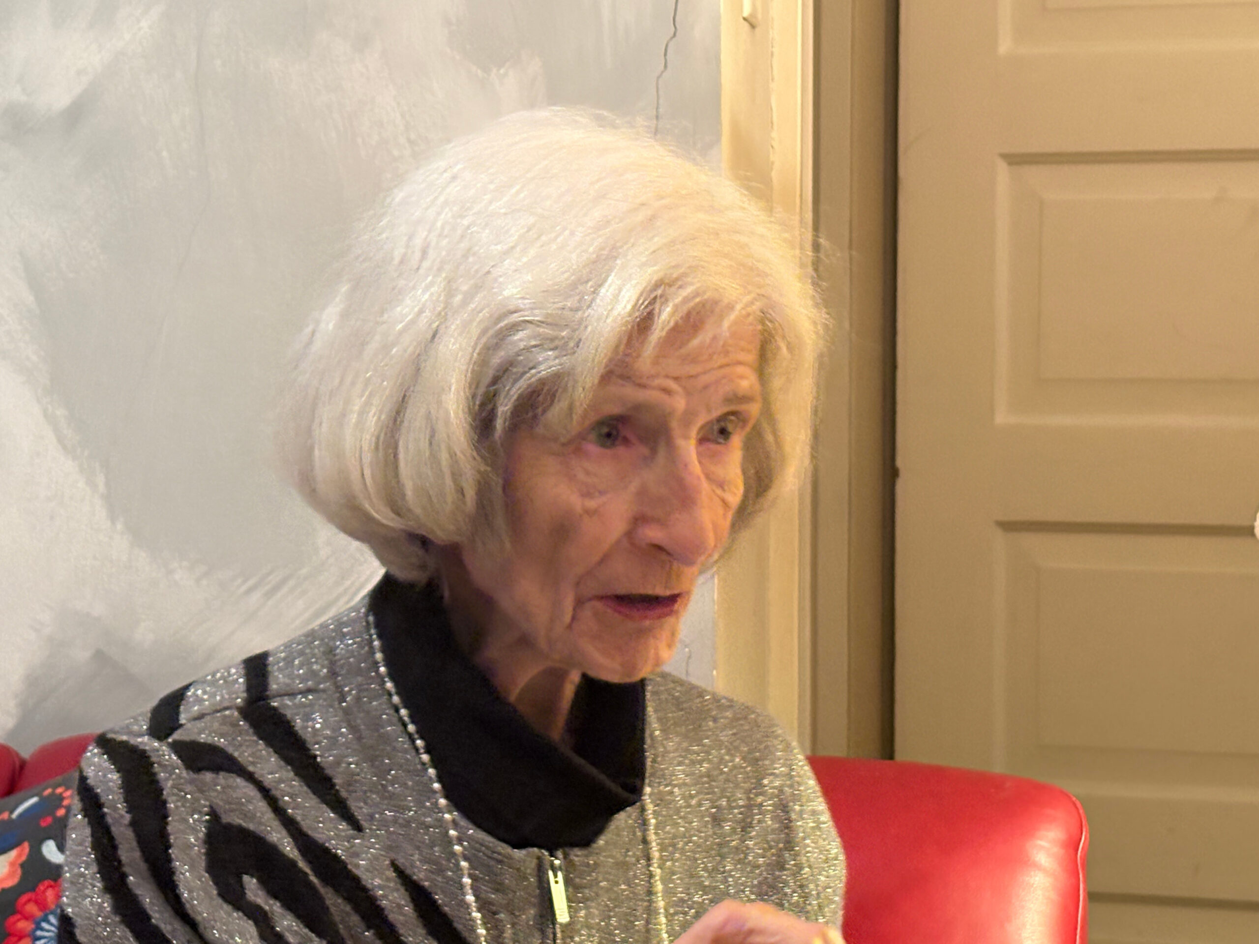 Older woman with short white hair wearing a sparkly gray cardigan and black top, seated on a red chair indoors near a door, looking to the side.