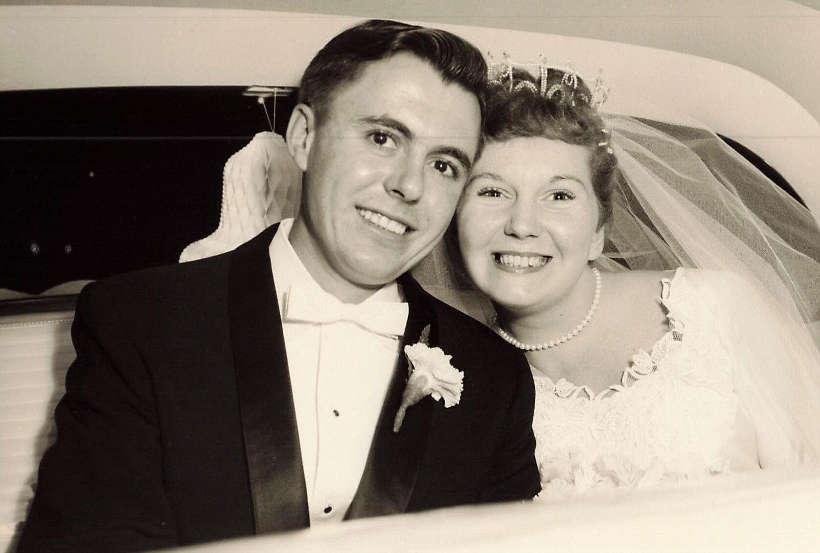 Bride and groom smile together in a vintage car on their wedding day, black-and-white photo.