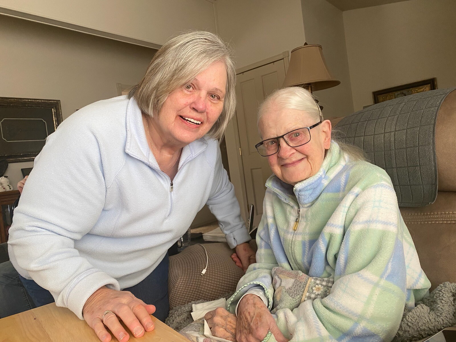 Two elderly women smiling at the camera in a cozy living room; one leans on a table while the other sits on a couch wrapped in a pastel plaid blanket.