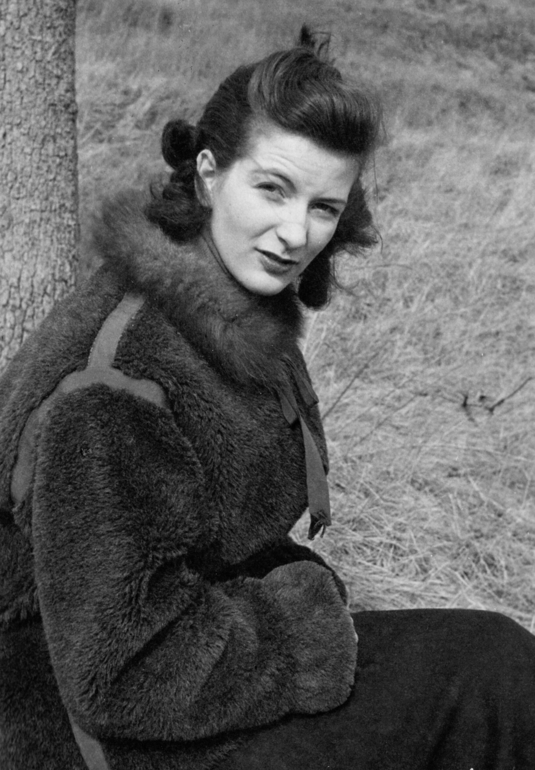 Black-and-white photo of a young woman in a thick fur coat sitting outdoors beside a tree, looking at the camera with a slight smile.