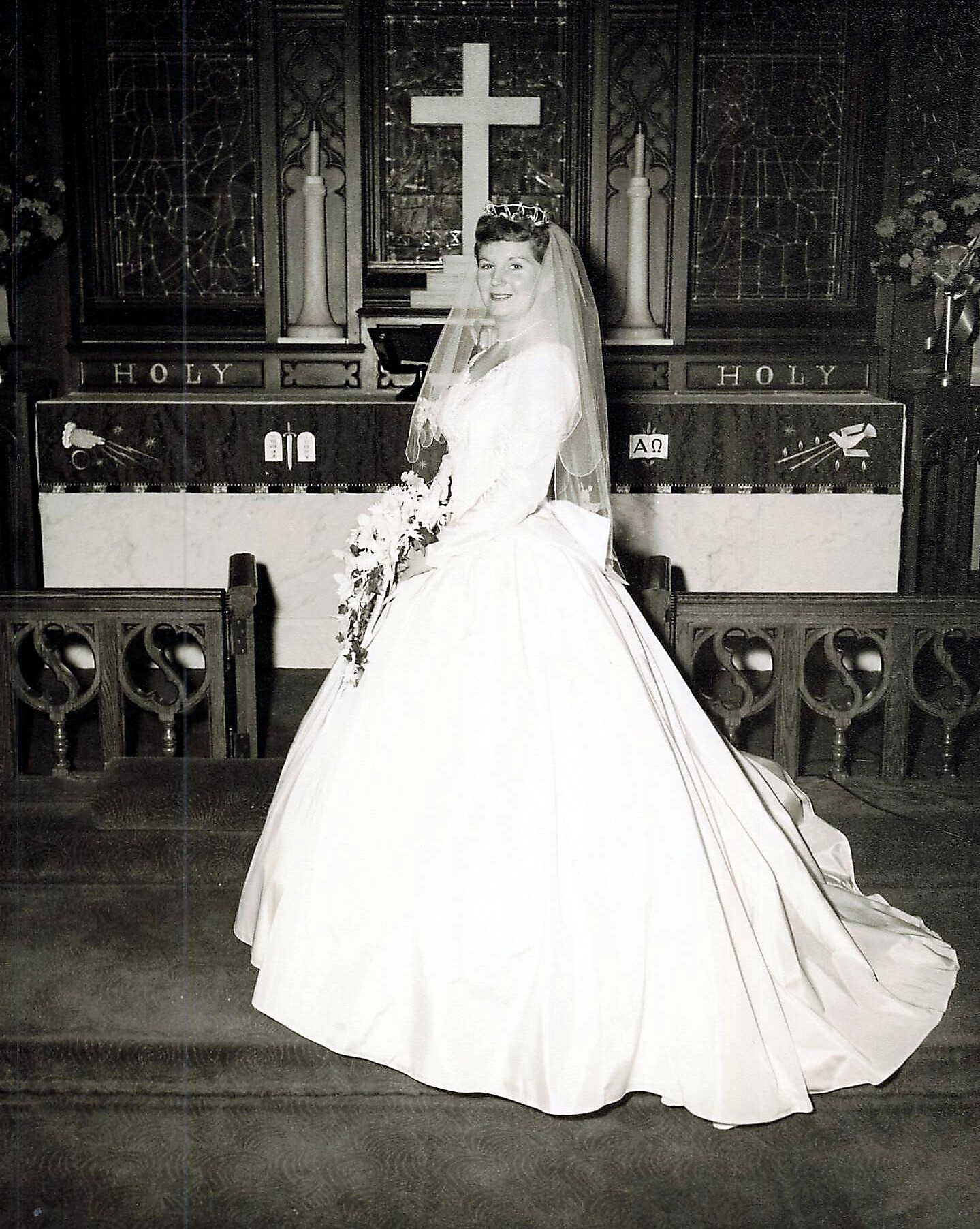 Bride in a wedding gown and veil stands at the church altar with a large cross behind her, smiling at the camera.