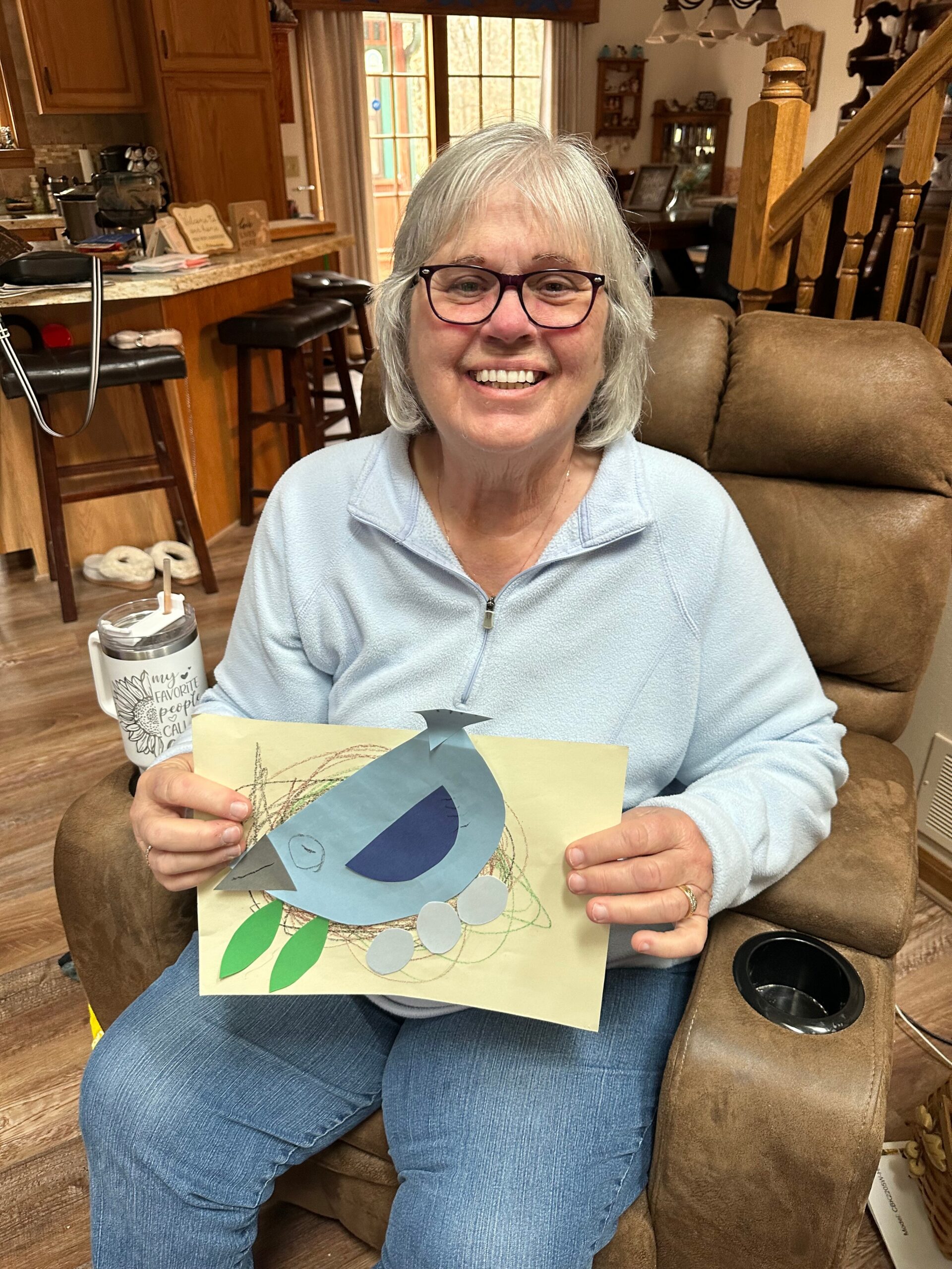 Smiling elderly woman with gray hair and glasses sits in a brown chair, holding handmade cards with paper shapes.