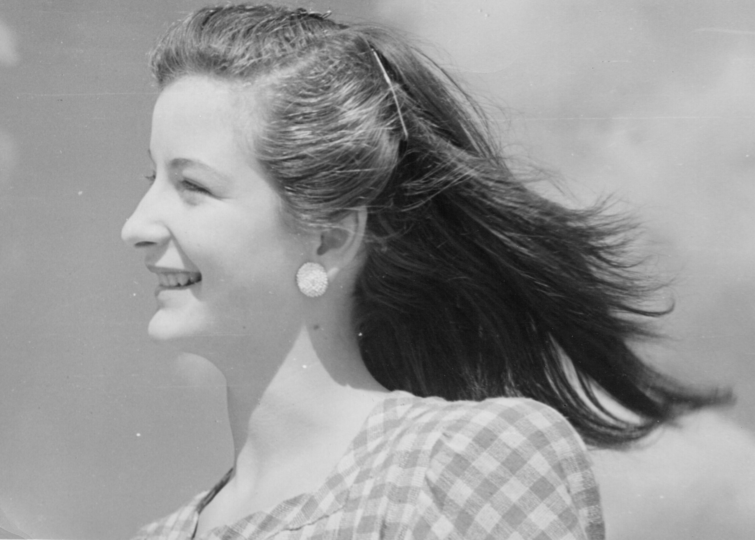Side profile of a smiling woman with wind-blown hair, wearing a plaid dress and round earrings.