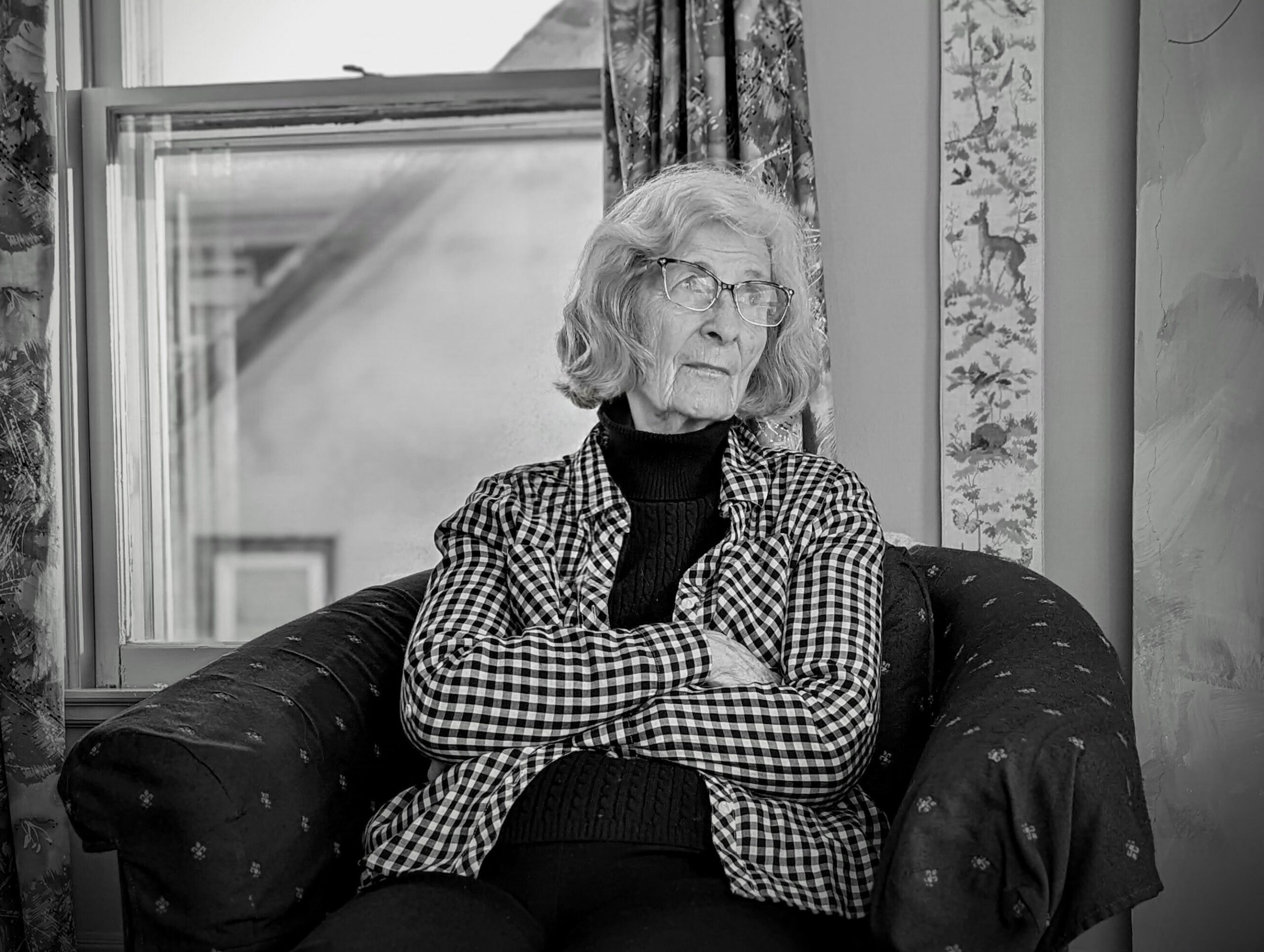 Older woman with gray hair and glasses sits in a patterned armchair by a window, arms crossed, wearing a checkered shirt and dark turtleneck.