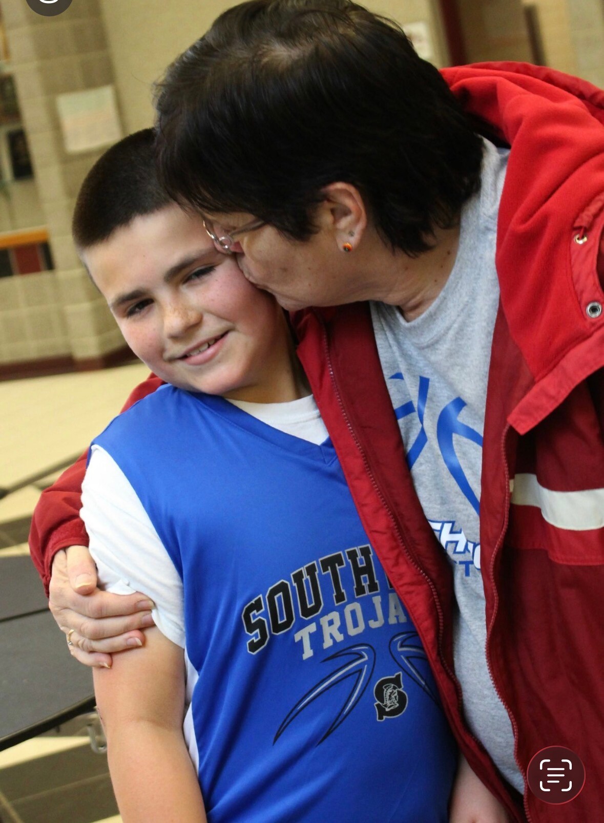 Two people embrace in a school hallway, with the older person kissing the younger boy on the cheek as they hug. The boy in blue smiles gratefully, wearing a blue athletic shirt.