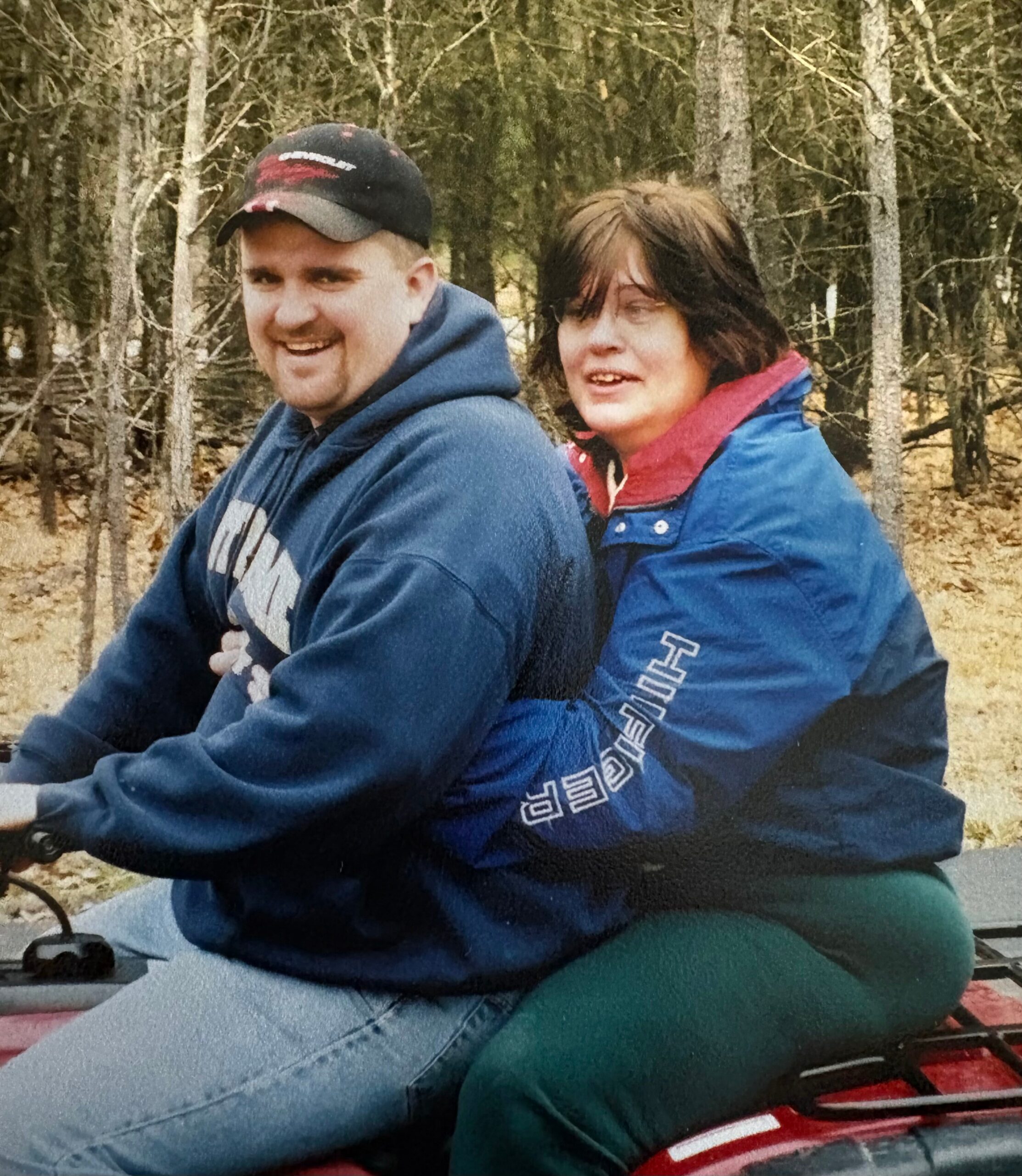 Two people sit close together in a wooded outdoor setting, smiling, wearing blue jackets and a red scarf in autumn leaves.
