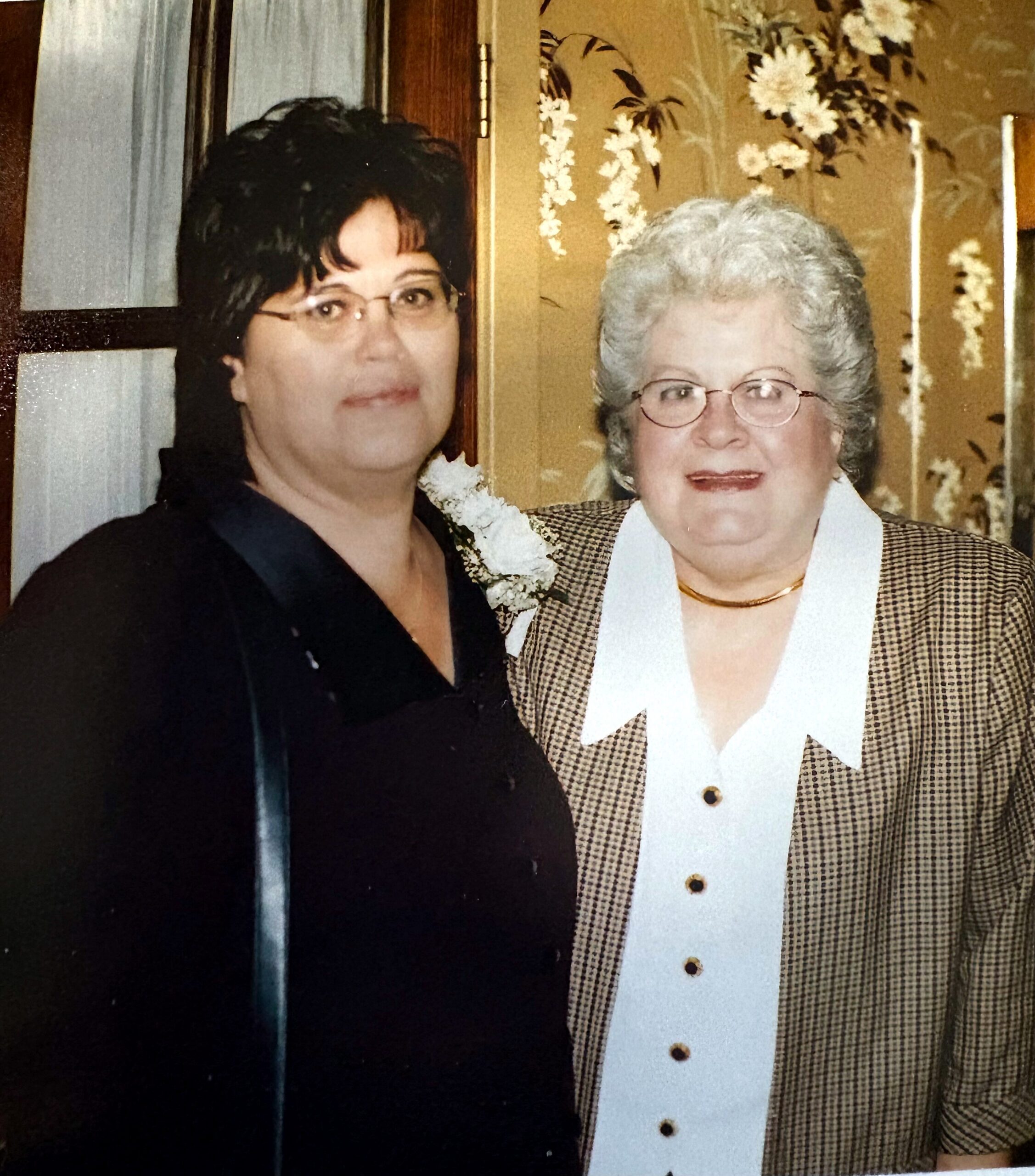 Two women posing for a photo indoors; one with dark hair and glasses on left, the other older with gray hair and glasses wearing a checkered jacket and white collar.