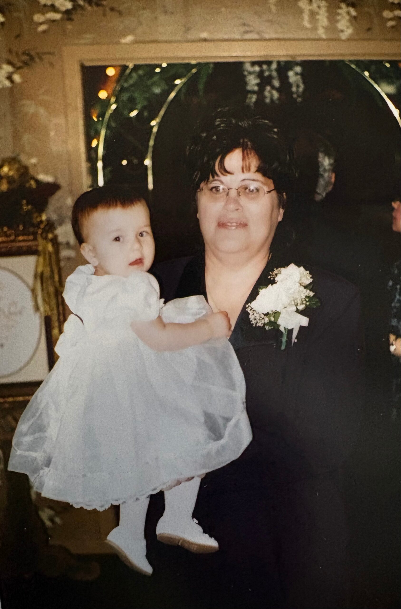 Older woman with short dark hair and glasses holding a baby girl in a white dress at a celebration; the woman wears a corsage.
