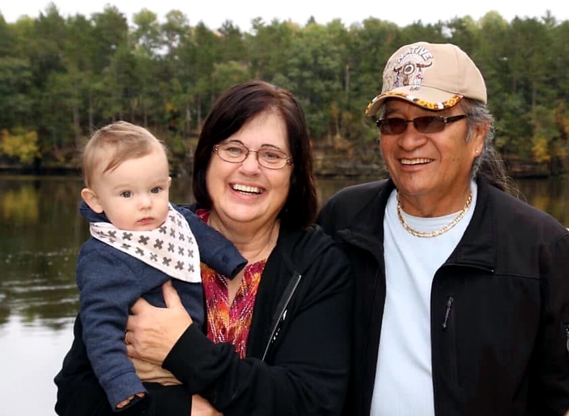 Smiling woman holds a baby while an older man stands beside them near a lake with trees in the background.