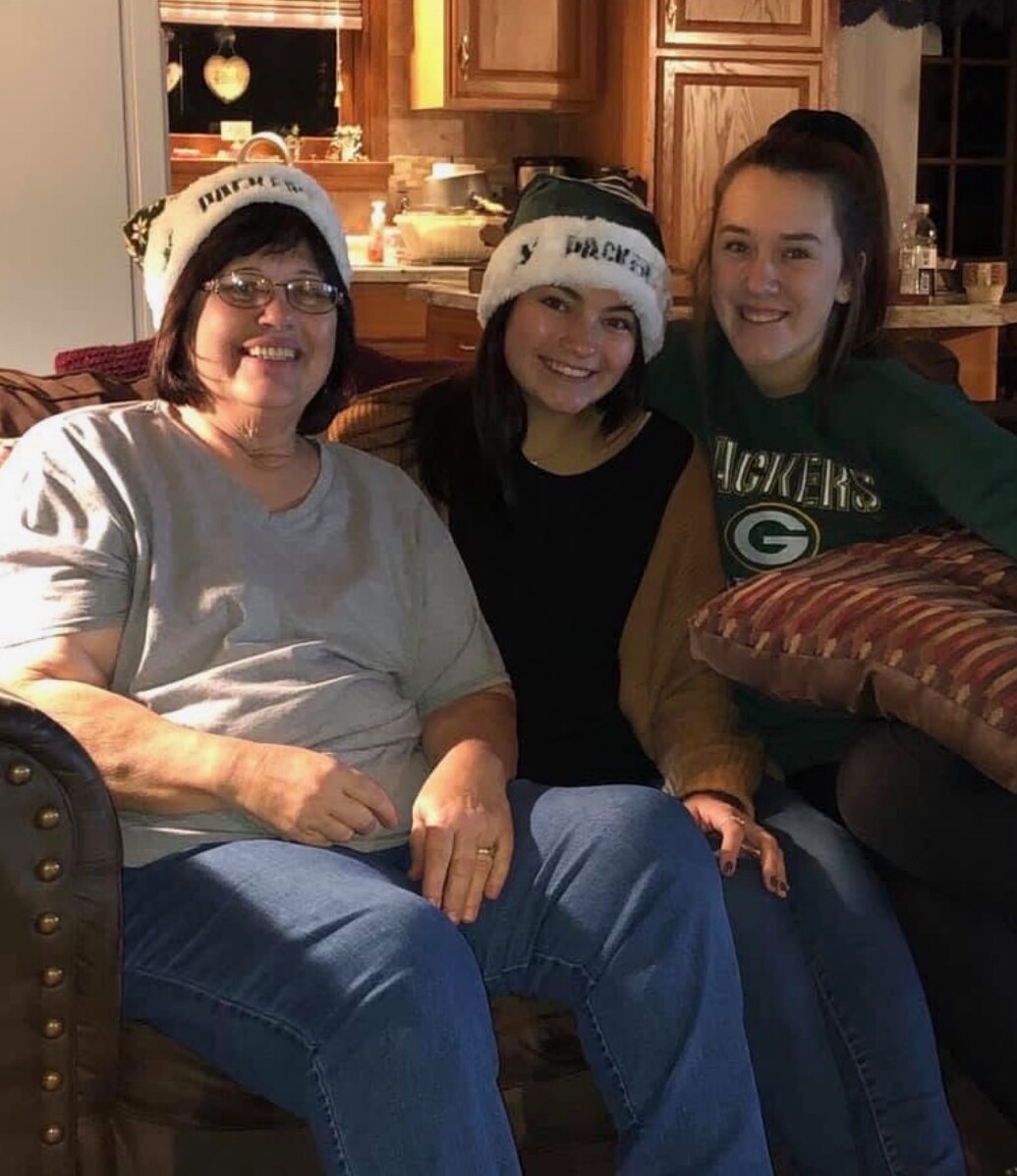 Three women sit on a brown couch in a cozy home, smiling and wearing white Santa hats with text on them.