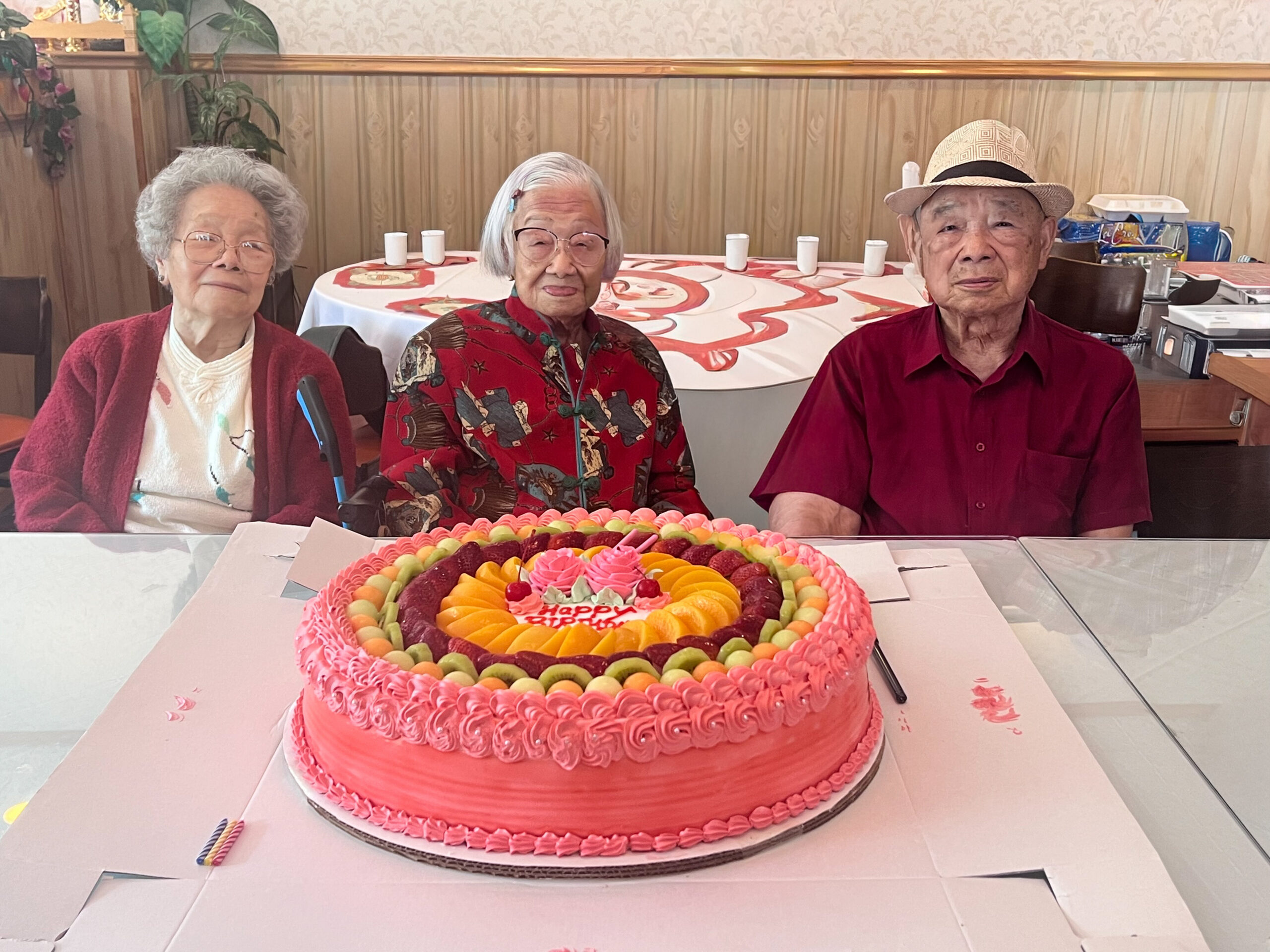 Three elderly people sit at a table behind a large pink frosted birthday cake with fruit toppings on display in front of them.