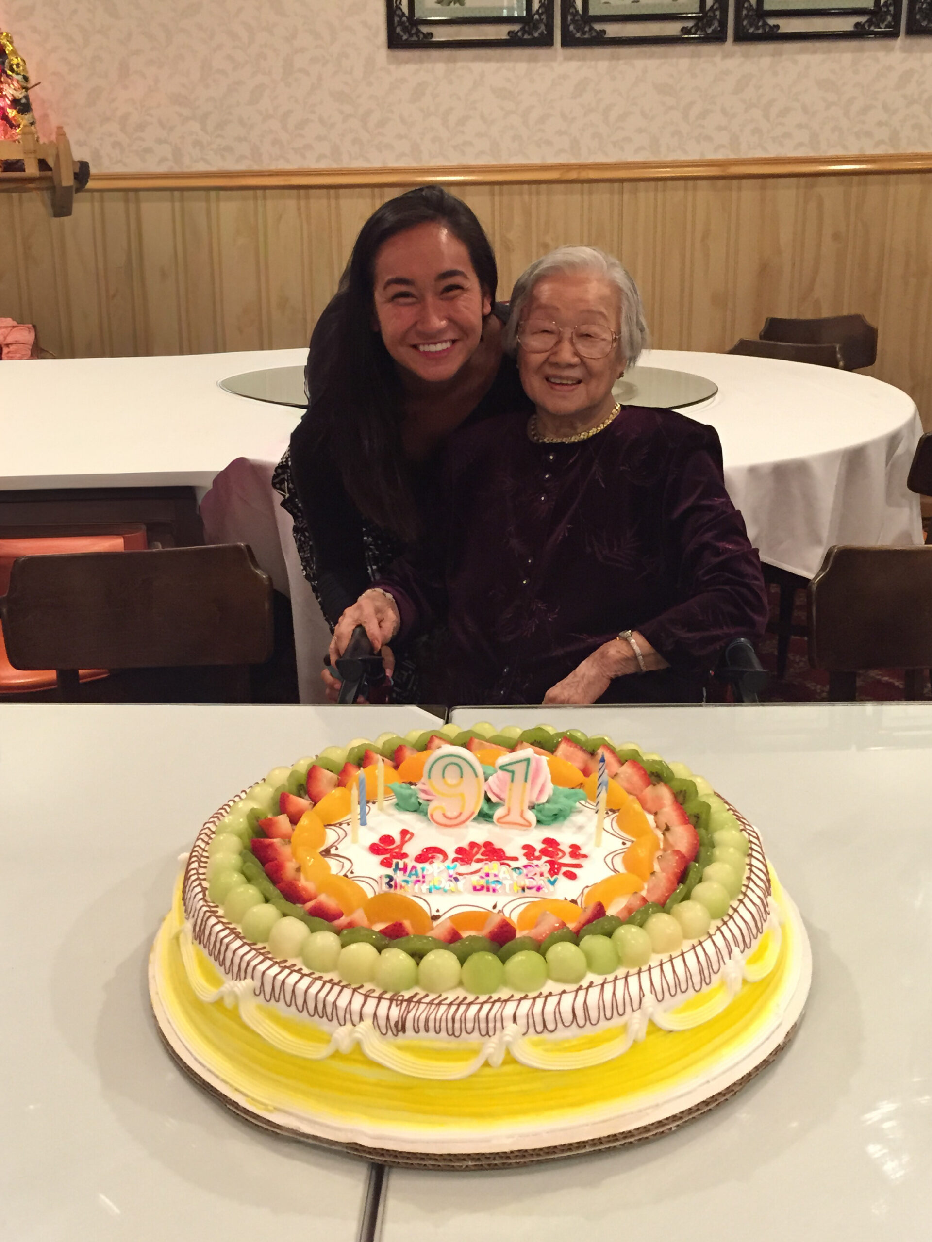Two women, one young and one elderly, smile as they pose at a birthday table with a large fruit-trost cake showing the number 91 (birthday cake).