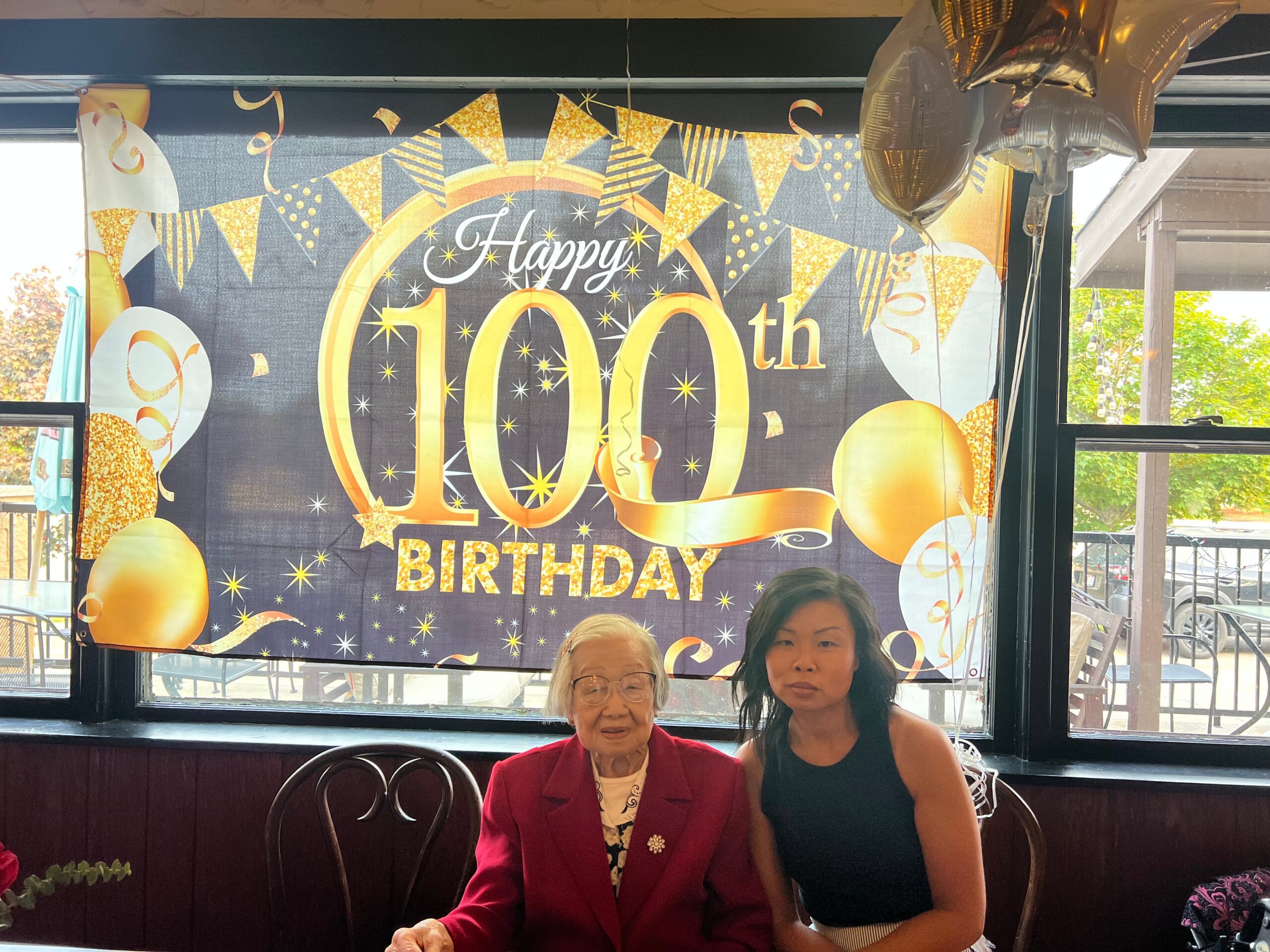 Two women sit at a table in front of a black-and-gold 100th birthday banner with balloons and confetti decorations behind them, celebrating a milestone.