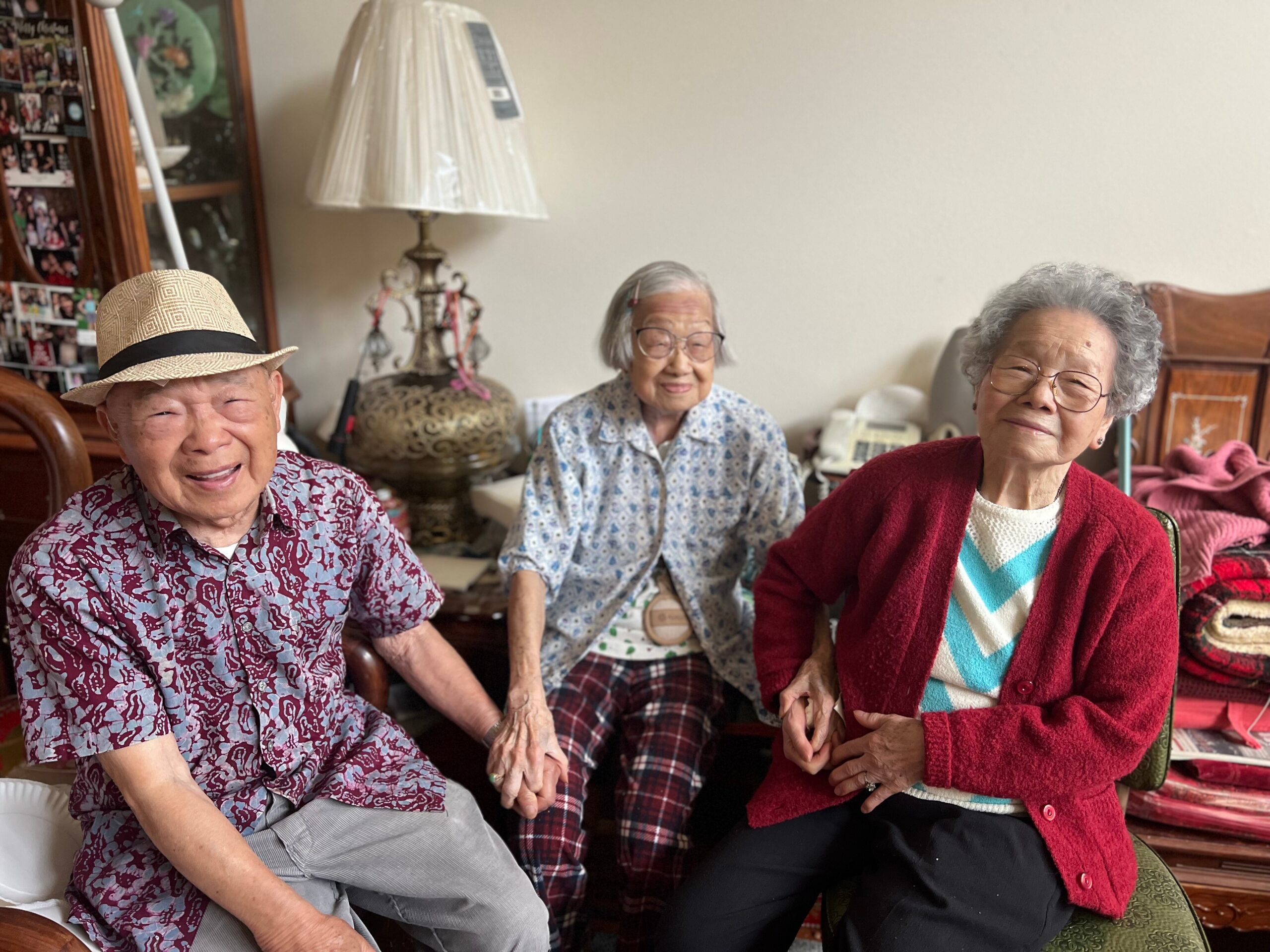 Three smiling seniors sit closely together in a cozy living room, wearing patterned clothing and looking at the camera.