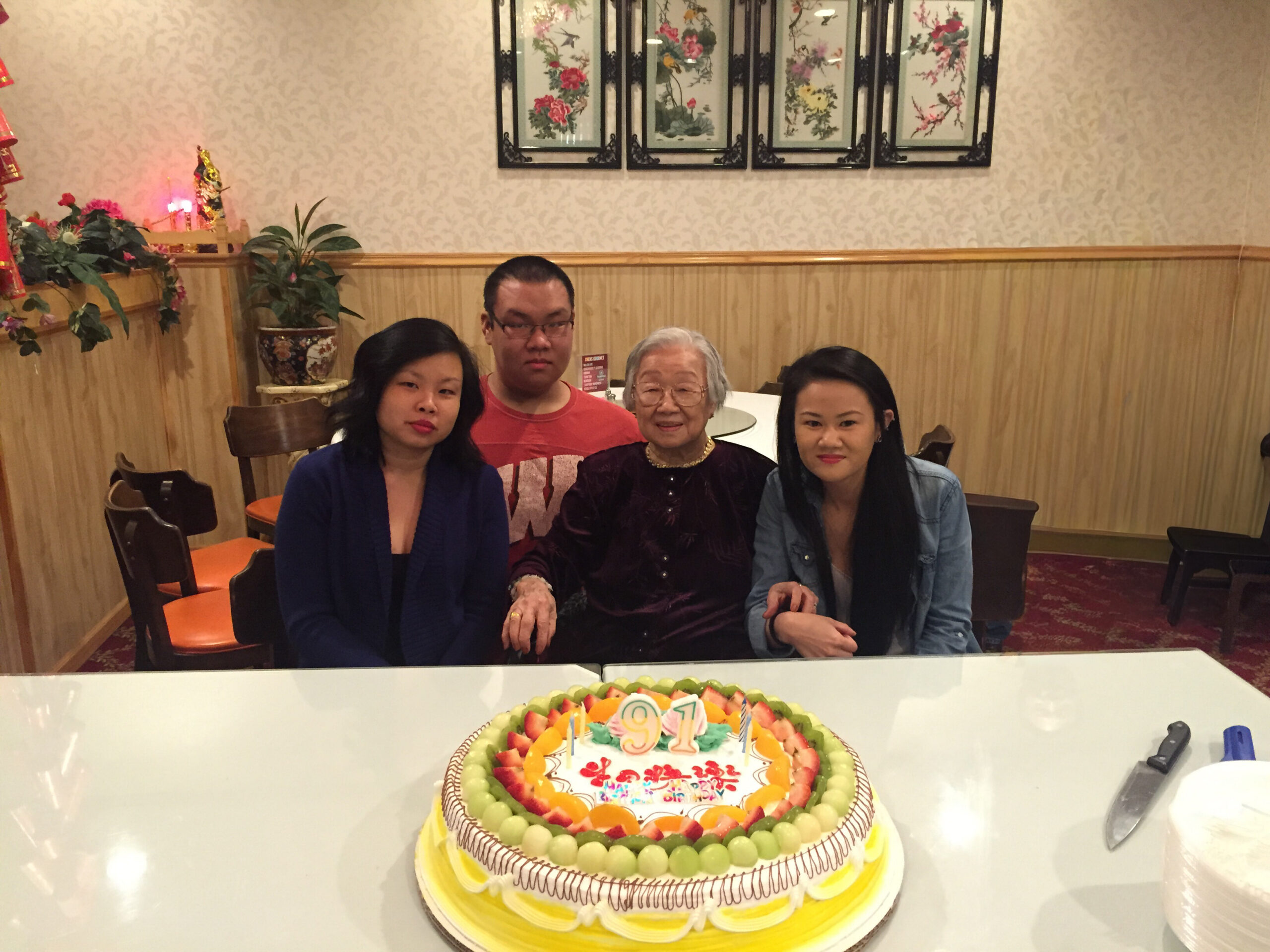 Four people sit at a table behind a large fruit-topped birthday cake in a restaurant setting, posing for a photo.