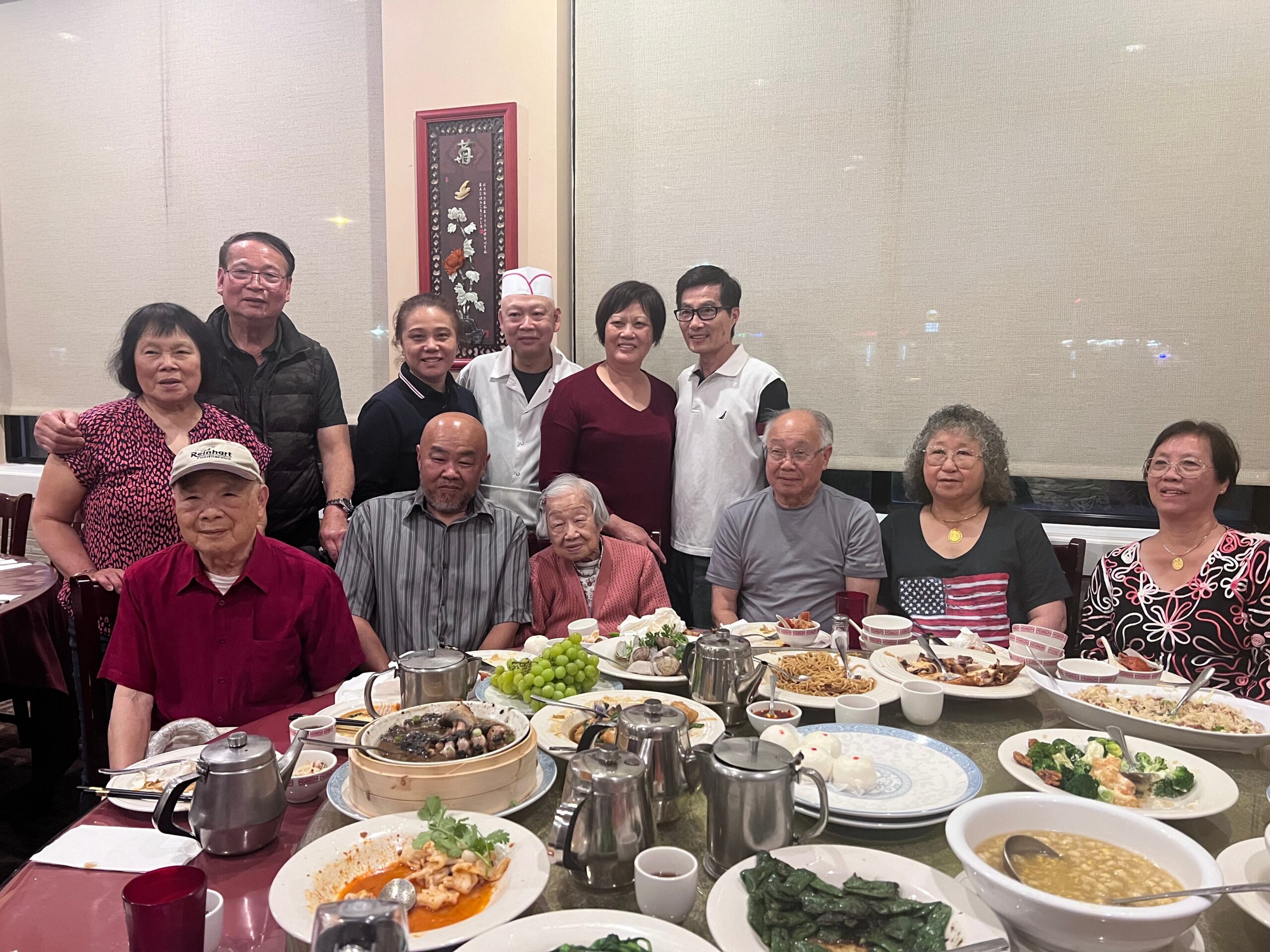 Group of mostly elderly Asian adults posing for a photo around a large banquet table with various dishes and drinks on display in a restaurant.