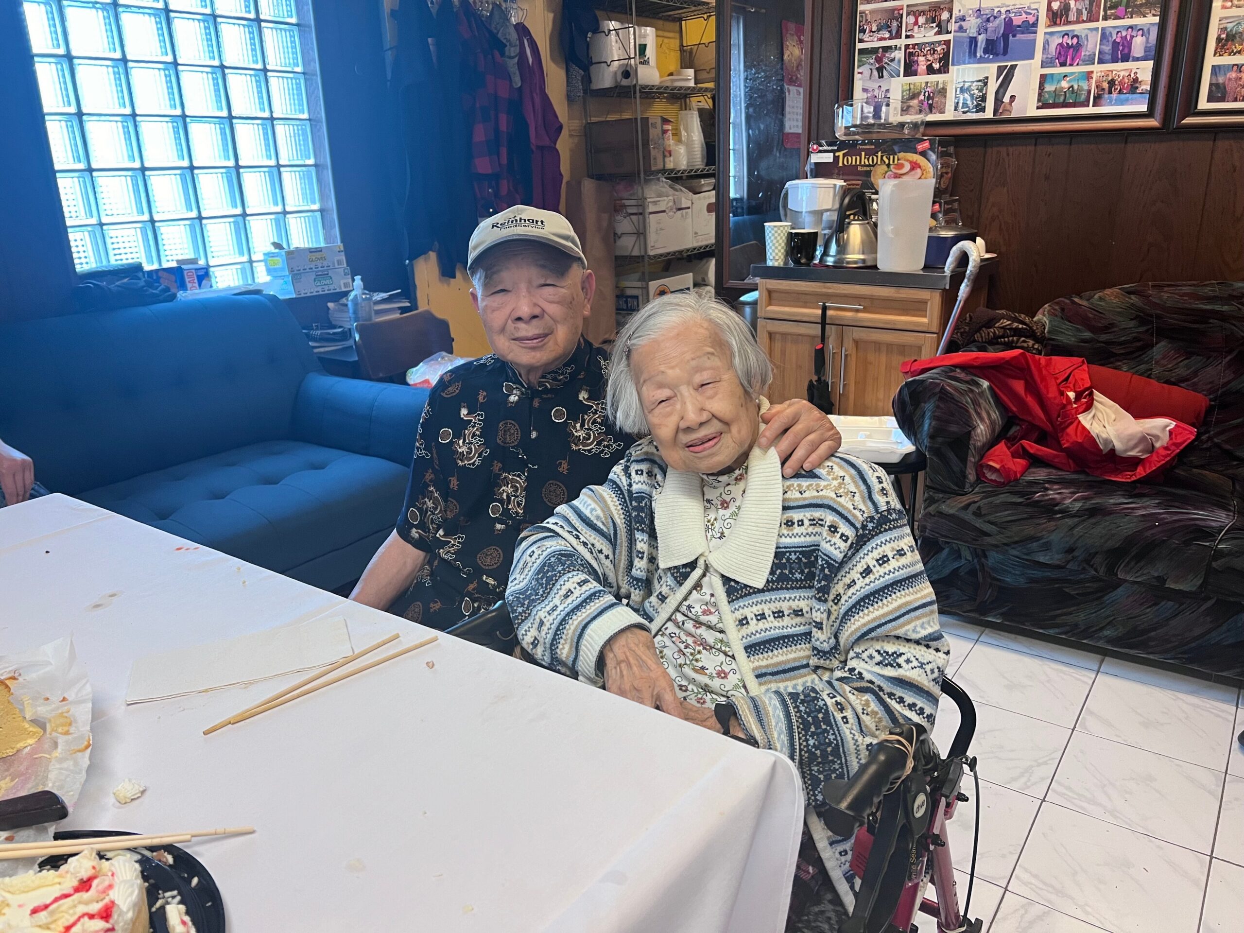 Two elderly people sit together at a table with a partially eaten cake and chopsticks, smiling at the camera in a cozy room with photos on the wall behind them.