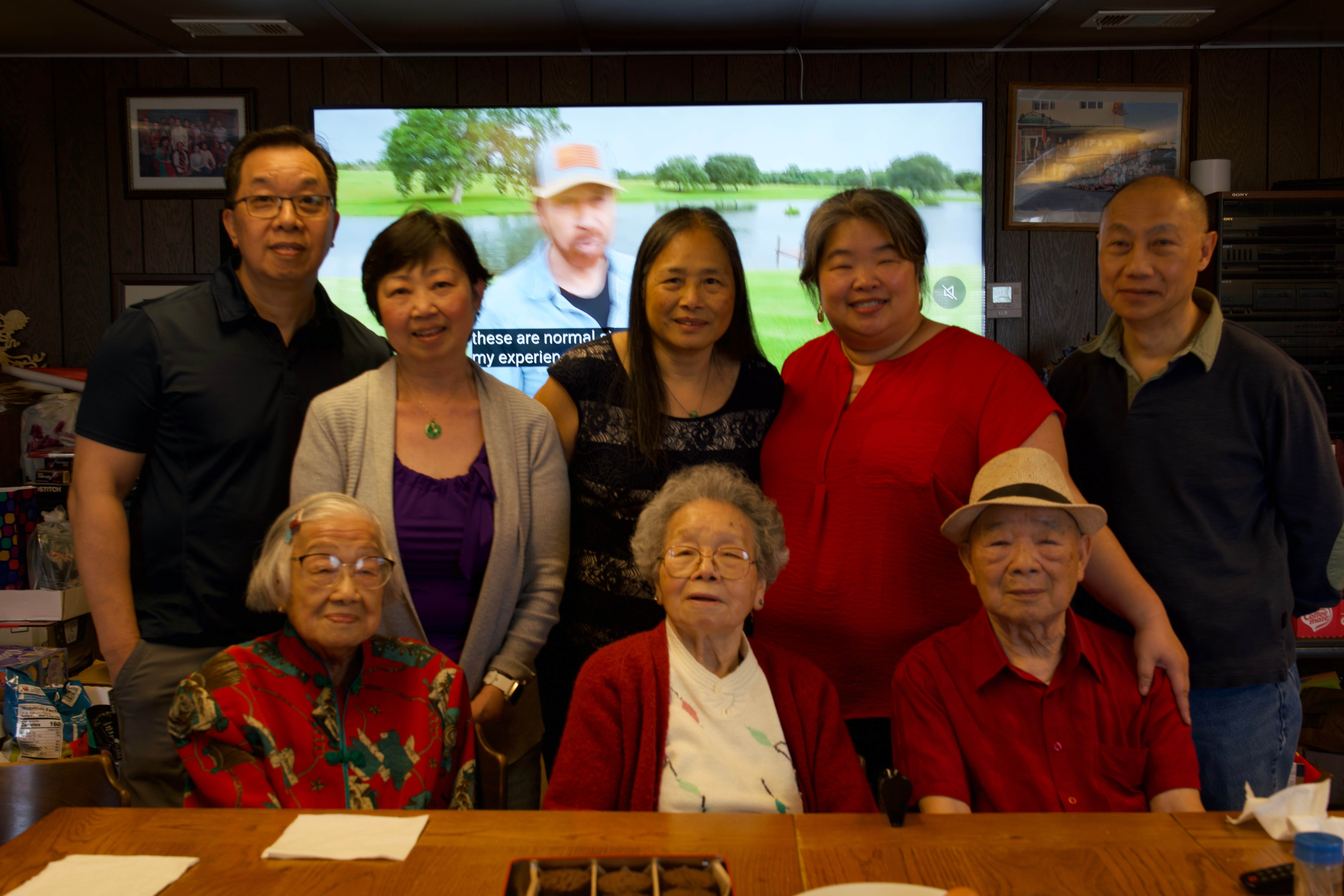 Group of adults posing for a photo around a table indoors, with a large screen showing a park in the background.