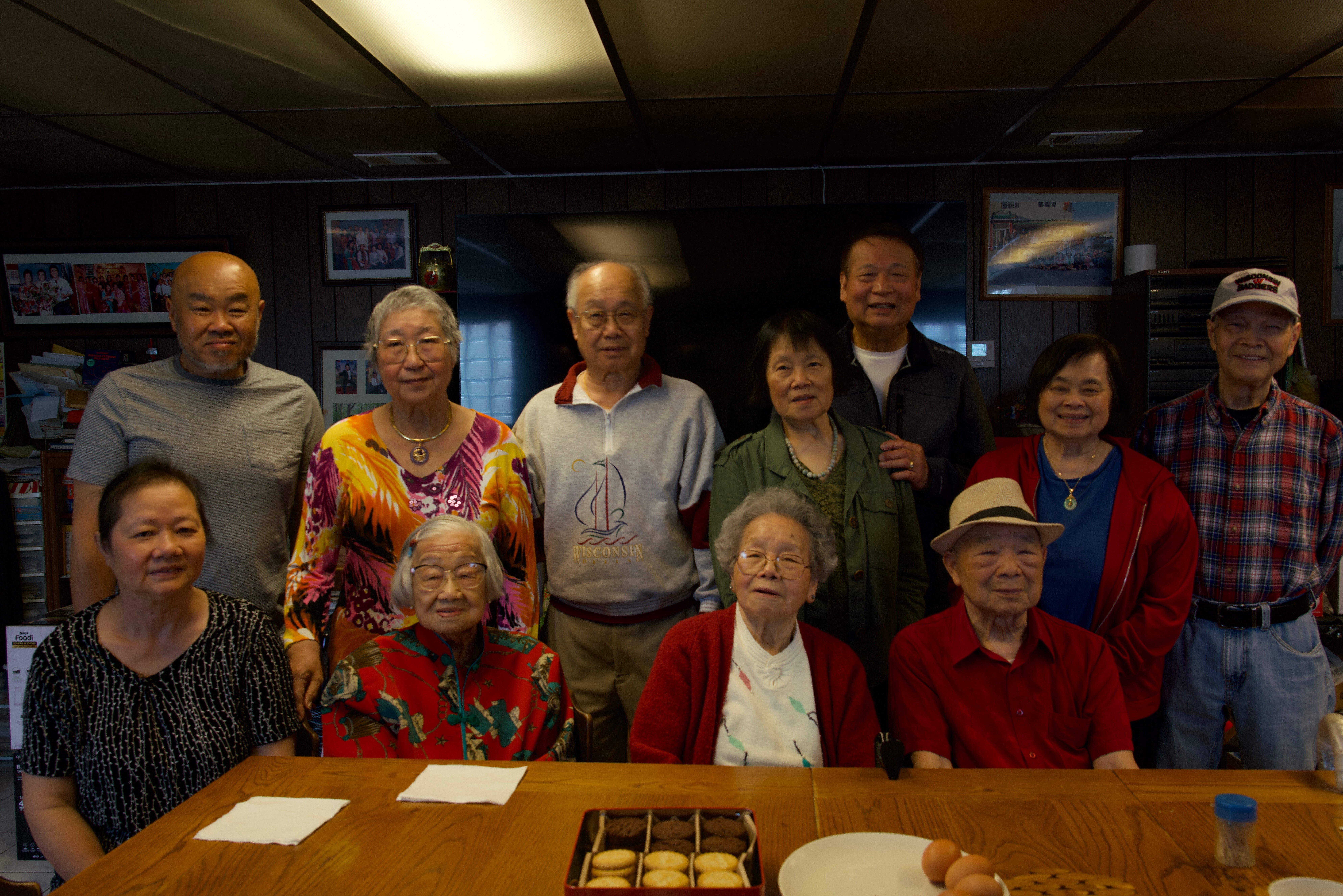 A group of seniors posing indoors in a wood-paneled room, with a tray of cookies on a wooden table in the foreground and eggs nearby, smiling at the camera.