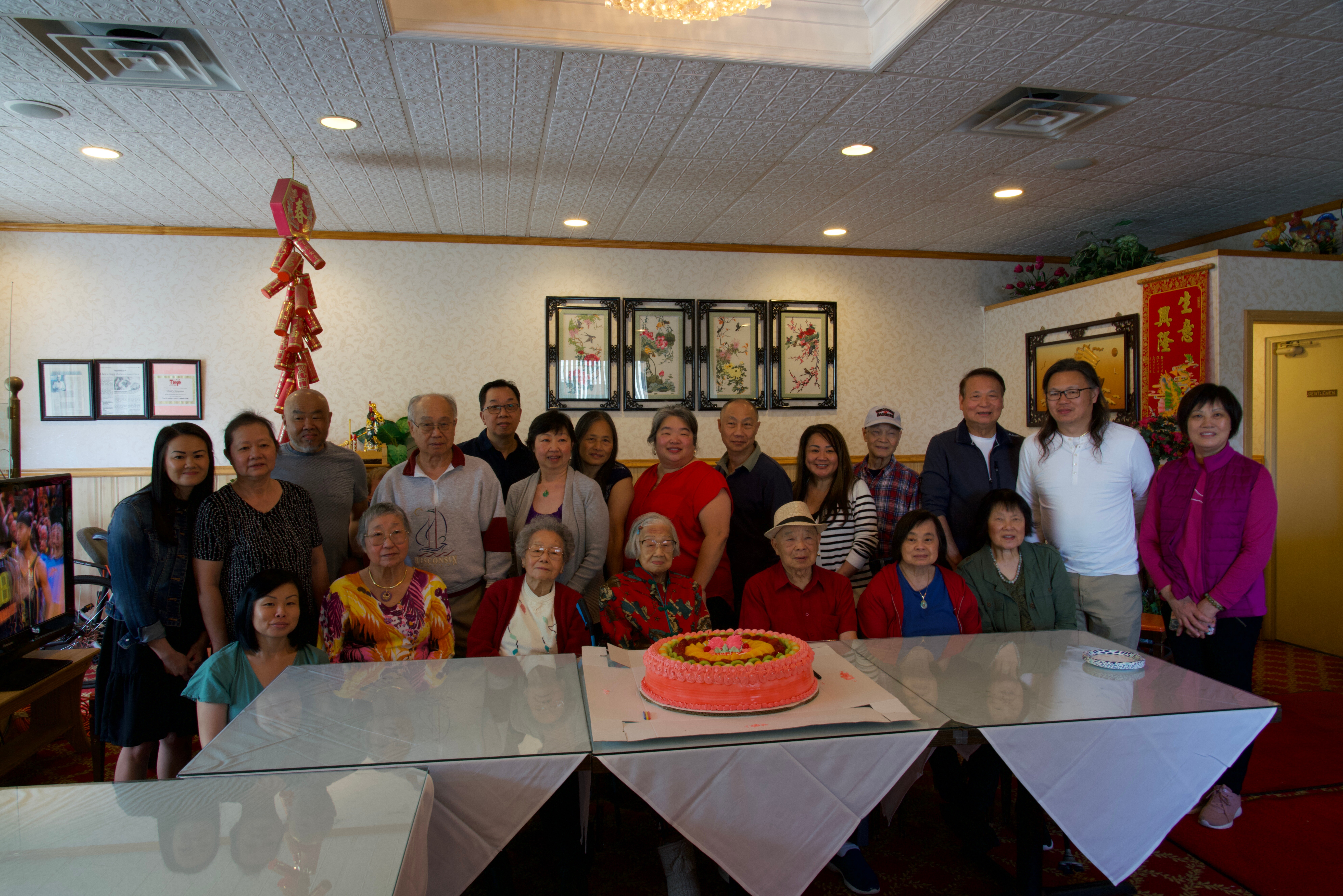 Group of people posing for a birthday-style celebration around a large pink frosted cake in a decorated dining room.