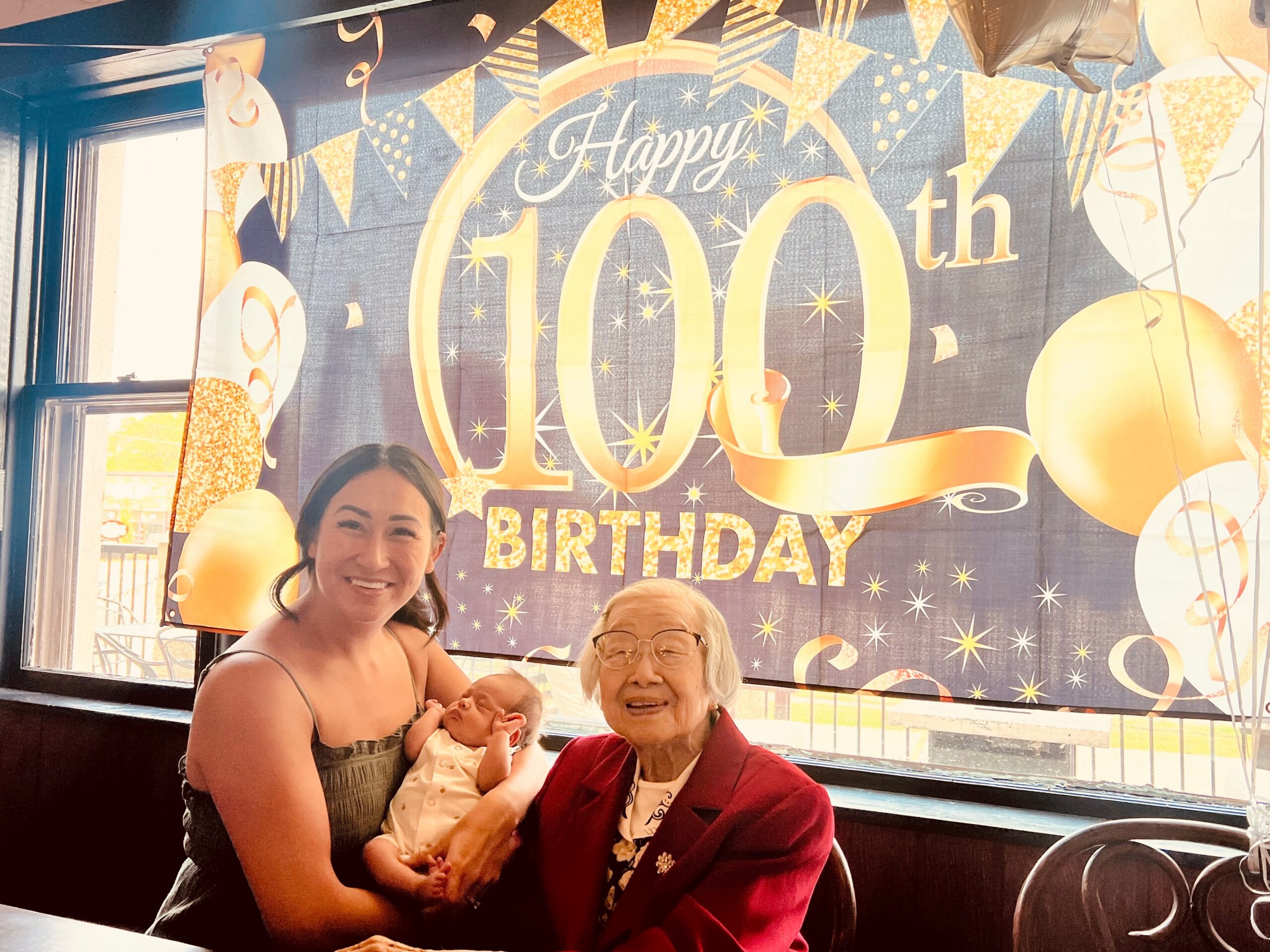 Smiling woman holds a newborn baby in front of a glittery 'Happy 100th Birthday' banner inside a bus. Two other people sit nearby, celebrating together.