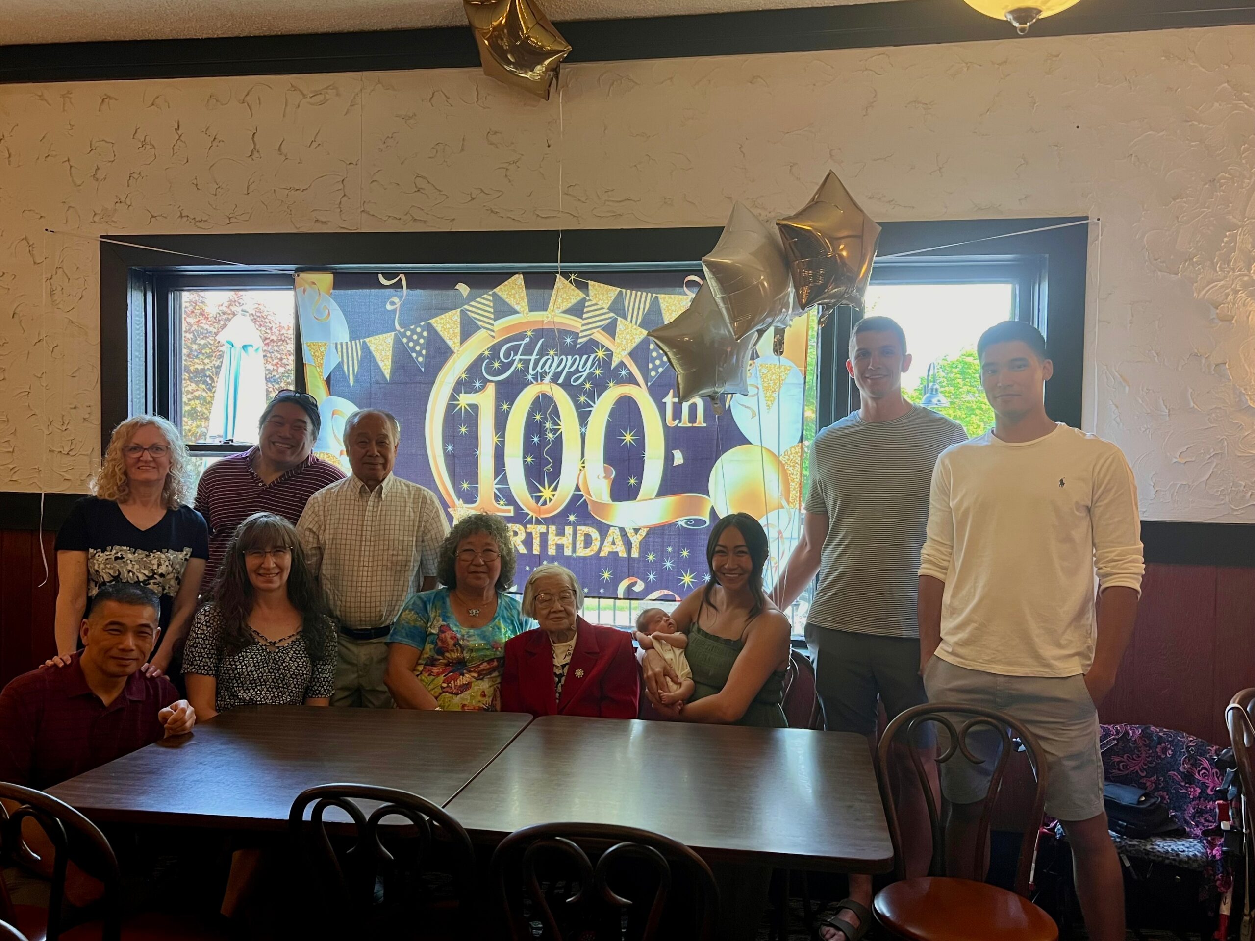 Group of adults and seniors posing indoors for a 100th birthday celebration, with a blue banner and gold balloons in the background.