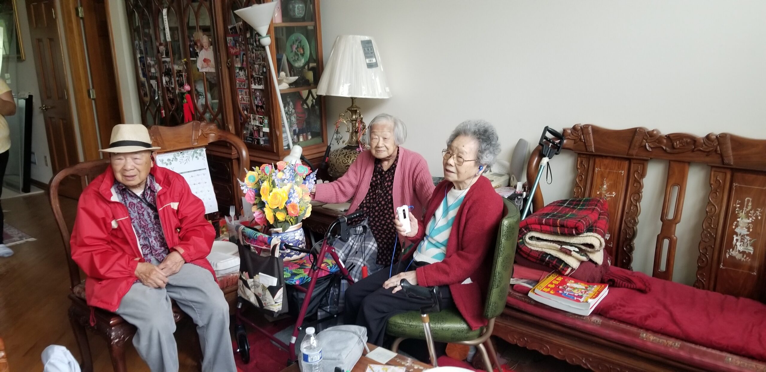 Three elderly people sit together in a living room, smiling, with a colorful bouquet on the table and a walker nearby.
