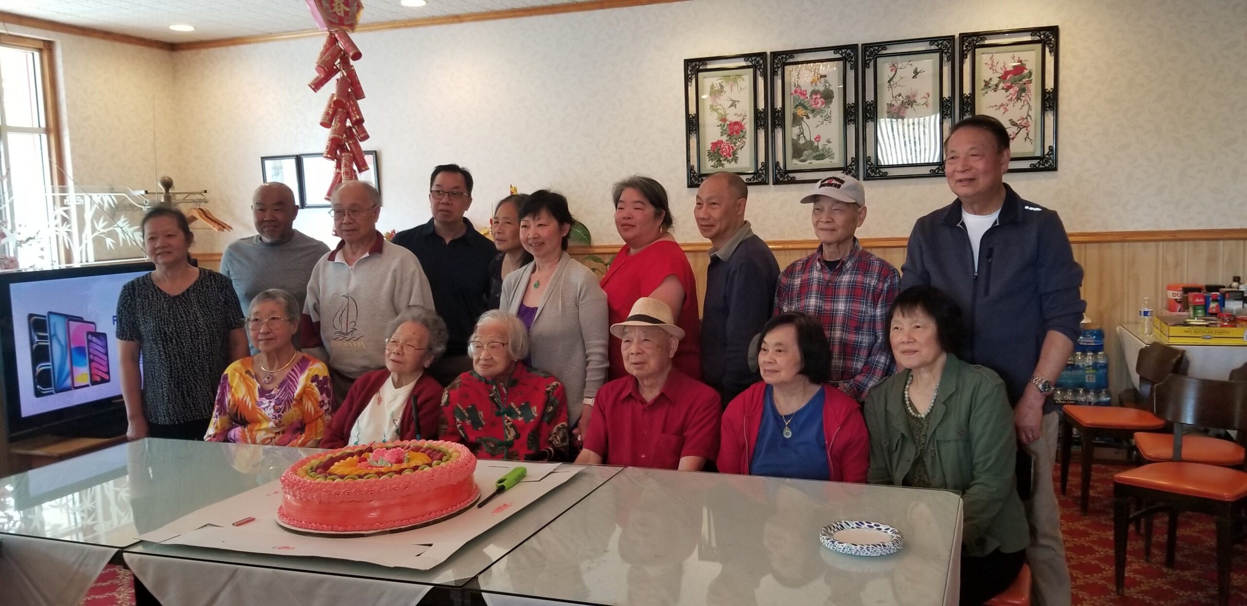 Group of elderly people posing for a birthday celebration around a large pink frosted cake on a table indoors.