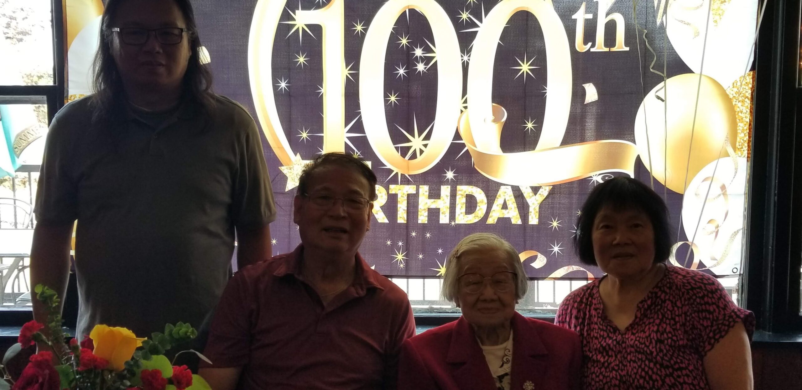 Four people (two men, two women) posing at a 100th birthday party, with a large '100th Birthday' banner in the background and flowers on the table.