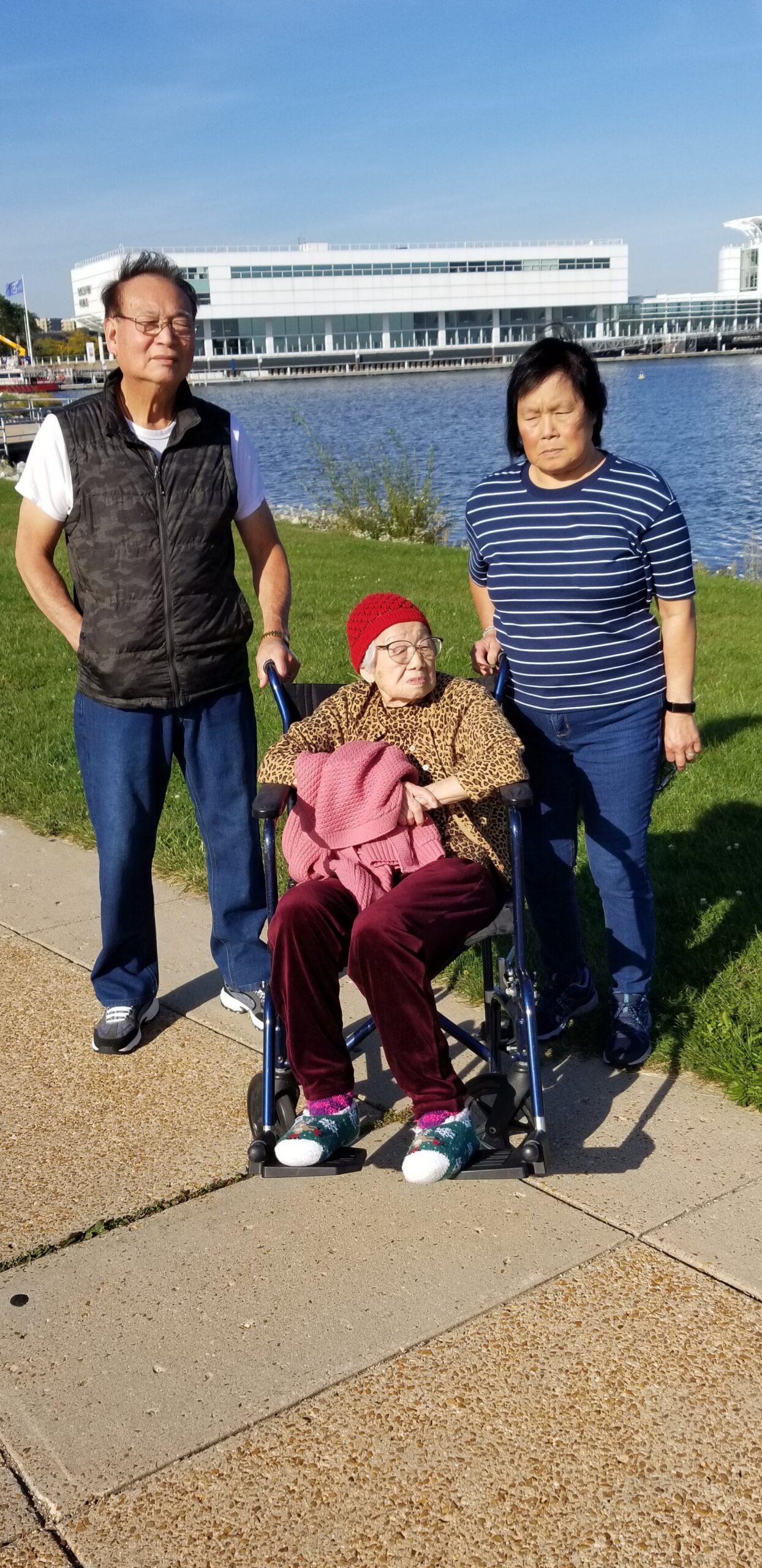 Three generations pose outdoors by a lake, with an elderly woman in a wheelchair flanked by a man and a woman.