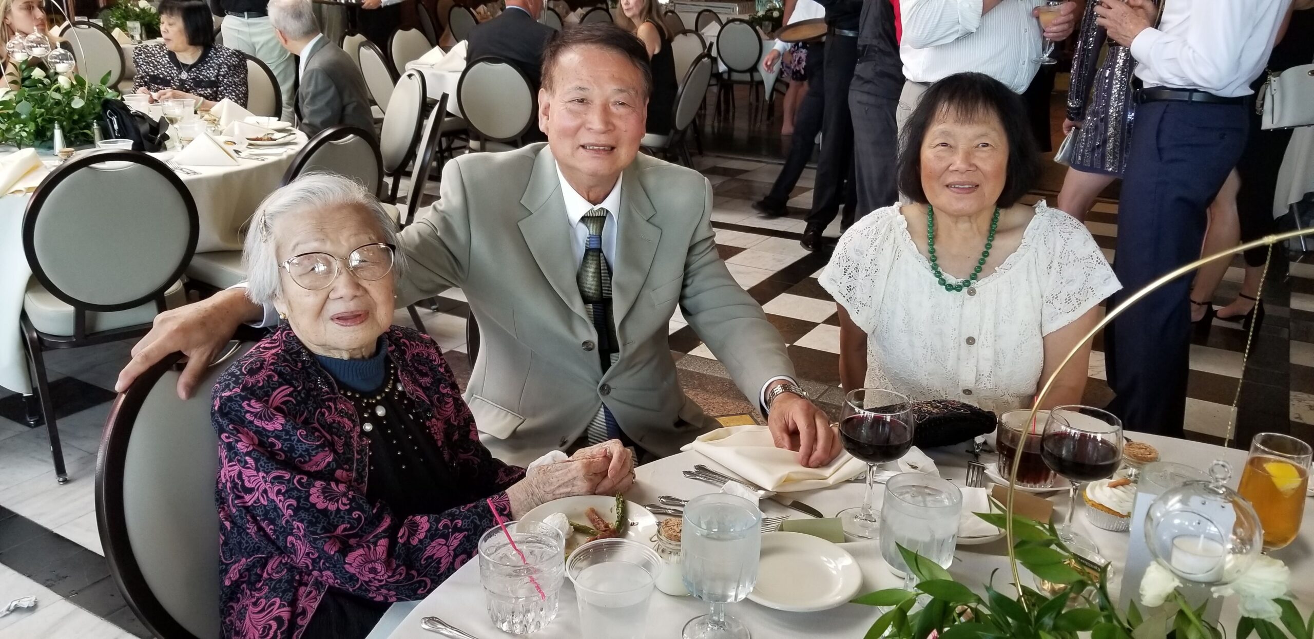 Three seniors pose together at a formal banquet table, smiling for the camera: an elderly woman with glasses on the left, a man in a gray suit in the center, and a woman with a green bead necklace on the right.