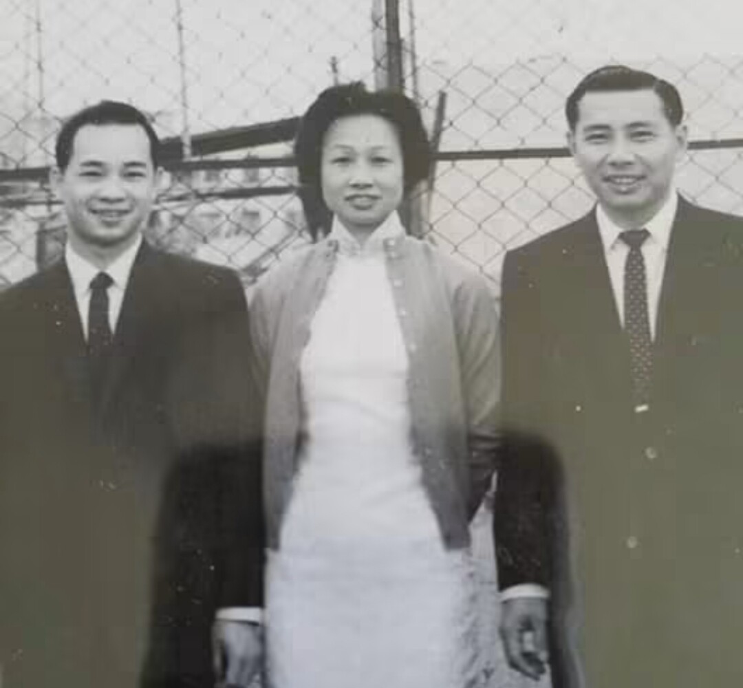 Three adults in formal attire posing outdoors in front of a chain-link fence: a woman in the center flanked by two men in suits.