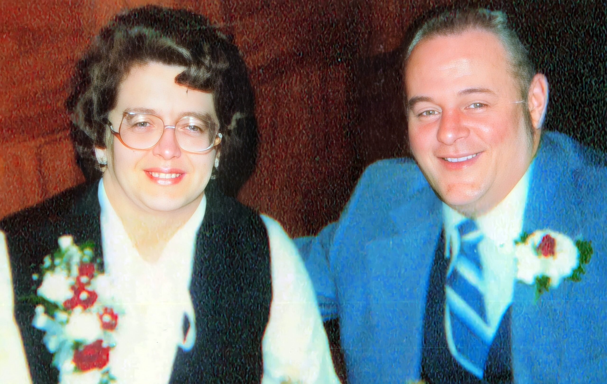 Two people in formal attire posing for a portrait, a woman with short dark hair and glasses next to a smiling man in a blue suit with a boutonniere on his lapel.