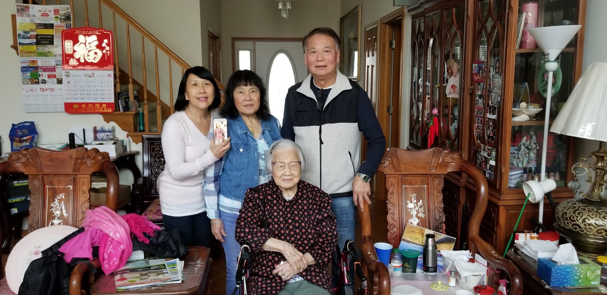 Family posing for a photo in a cluttered living room; an elderly woman sits in a chair while three relatives stand behind her, smiling.