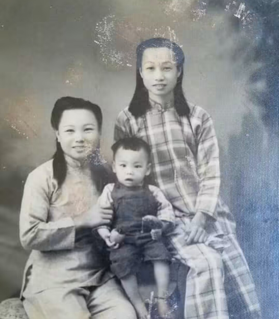 Black-and-white studio portrait of two women with a small child: a seated mother on the left holding her toddler, and another woman standing to the right in a plaid dress.