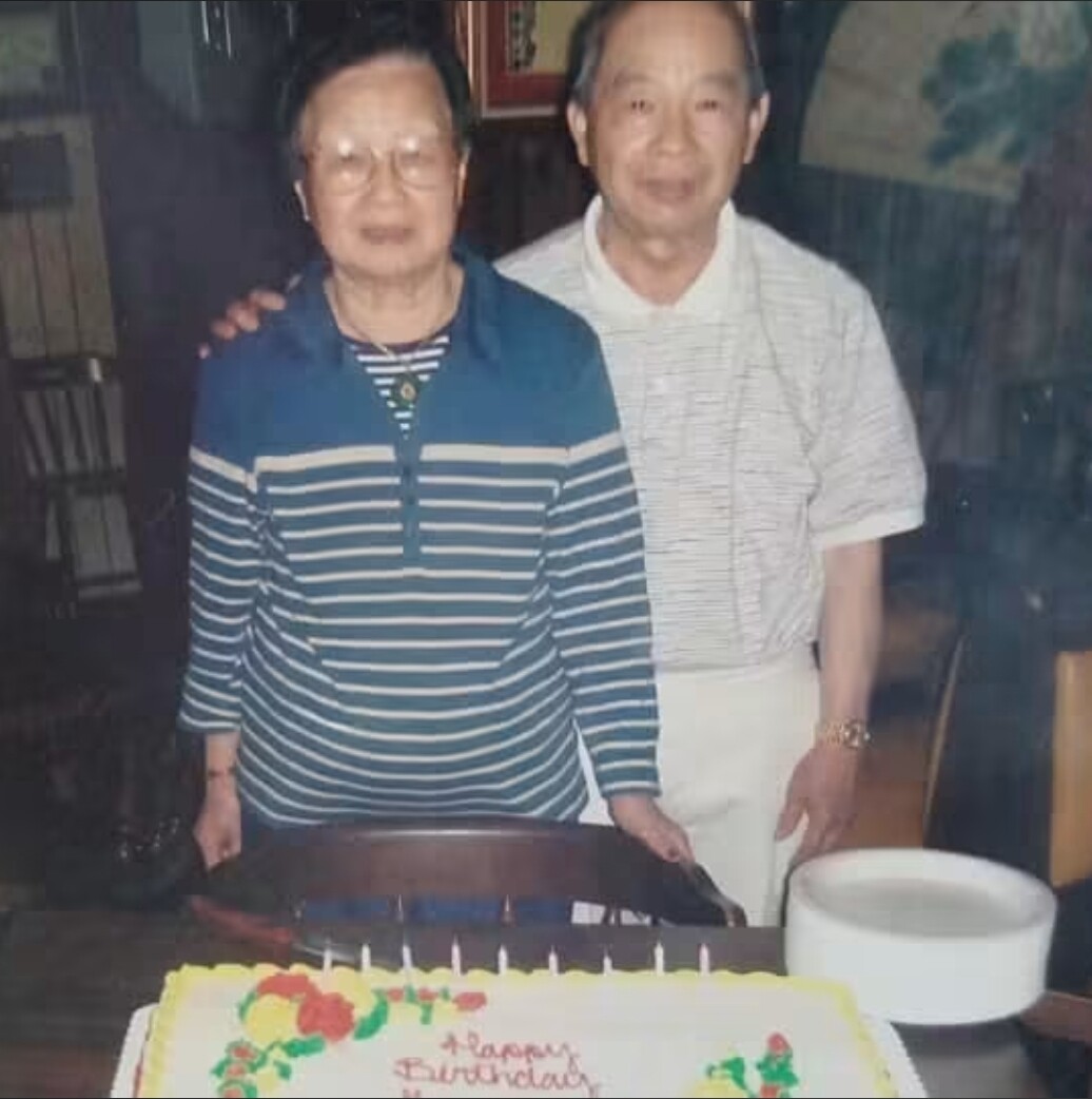 Two elderly people pose beside a rectangular birthday cake with lit candles on a table in an indoor setting.