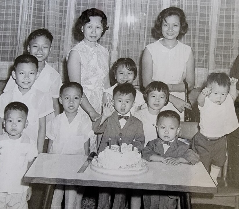 A birthday party with a cake on a table; a boy in a suit and bow tie sits by the cake while others stand around him, some waving.