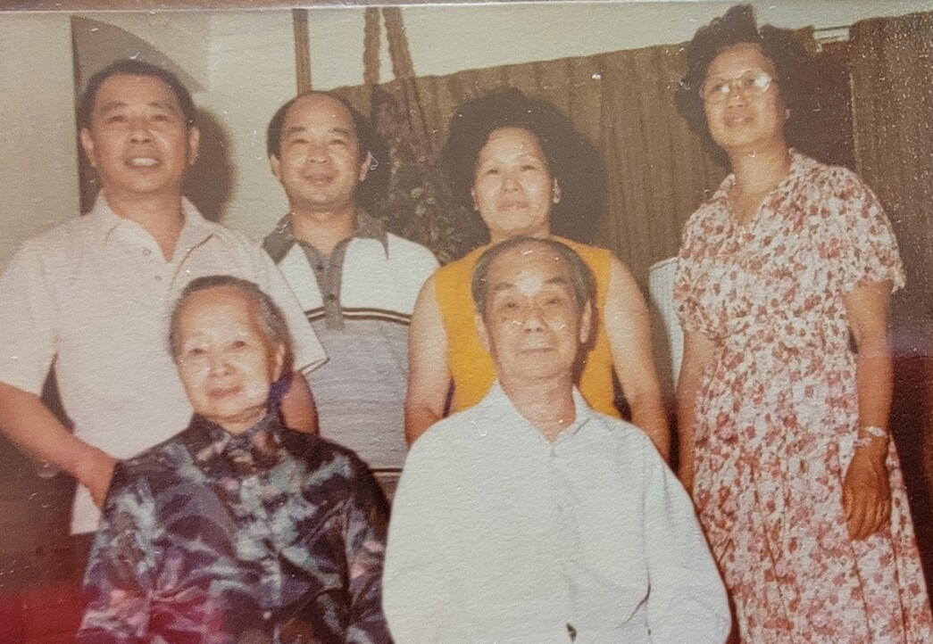 Six people posing for a family portrait indoors: two elderly seated in front, four adults standing behind them and smiling.