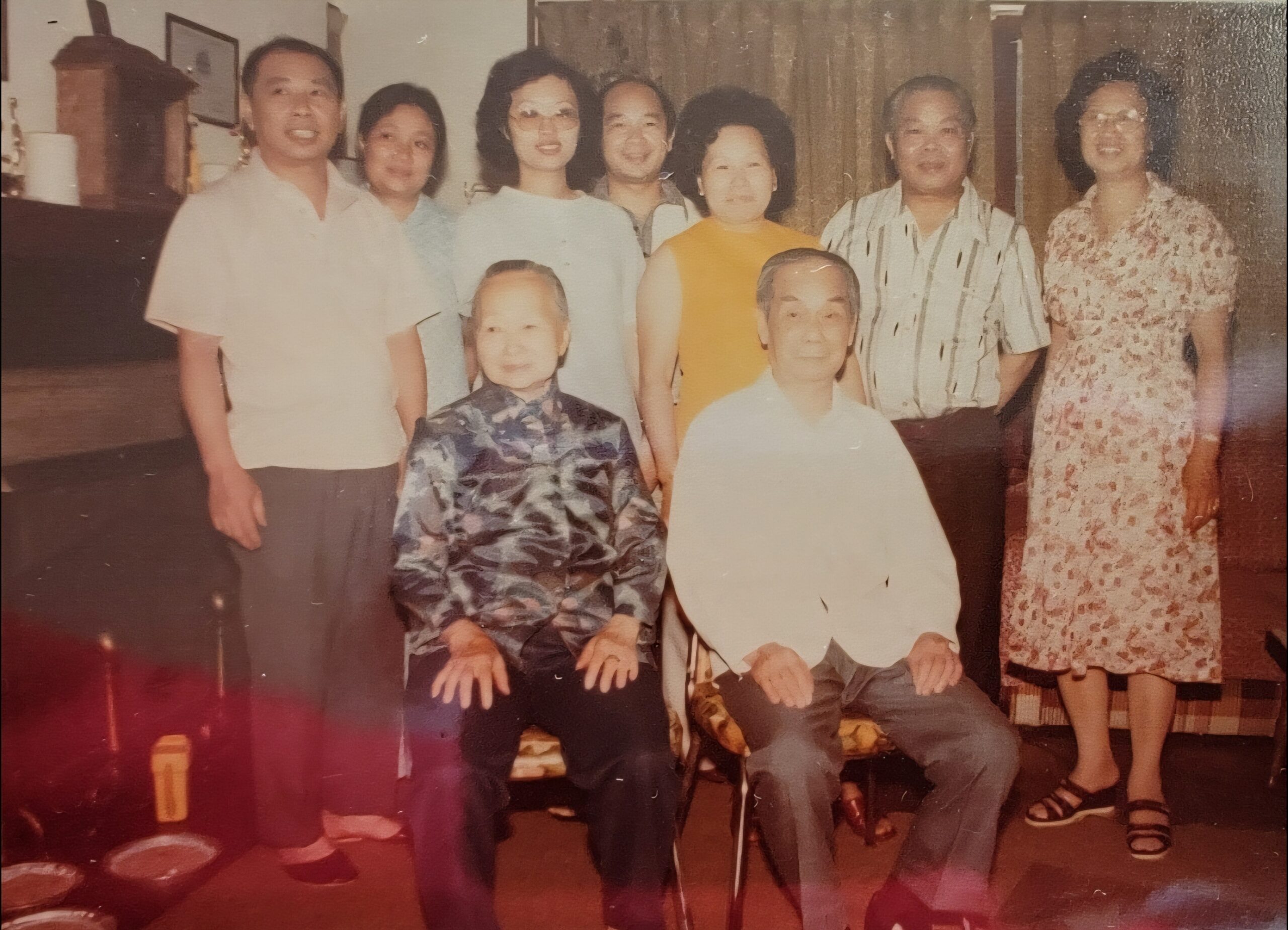 A multi-generational family group poses for a photo in a living room, eight adults standing behind two children seated in front, casual 1990s attire.