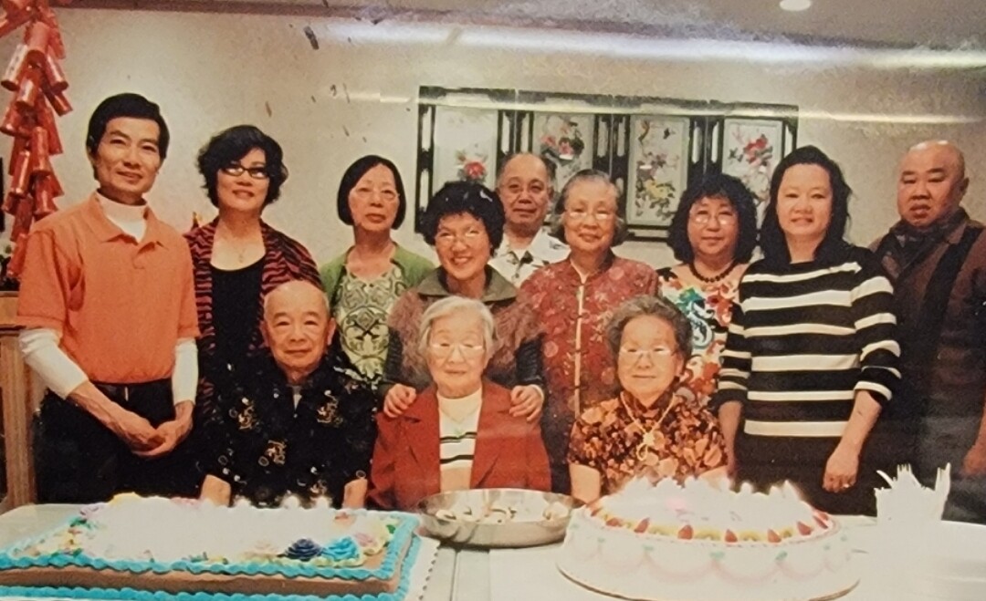 Group of predominantly elderly Asian people posing for a photo at a celebration, with two large decorated cakes in front and candles lit on one cake.
